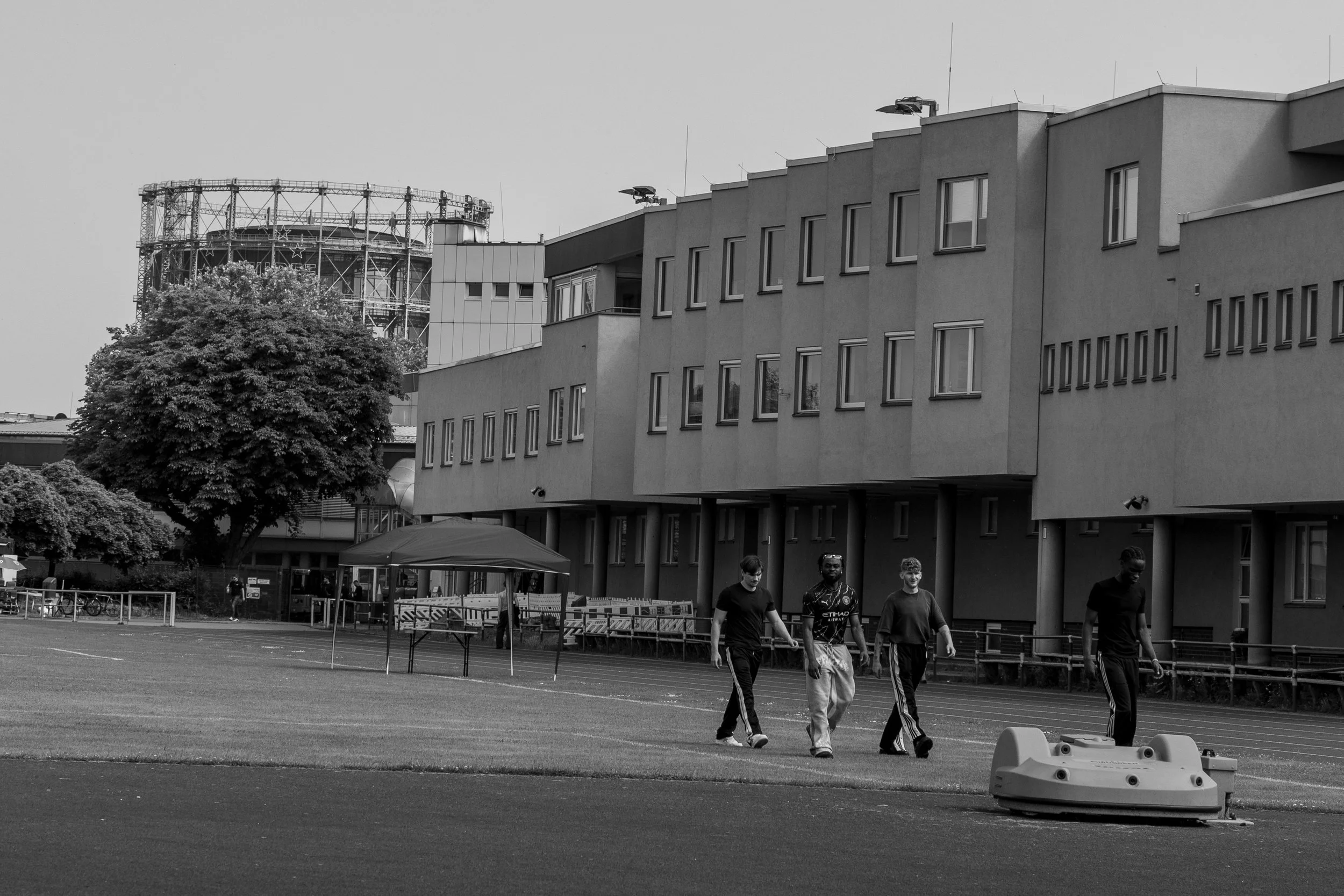 Schwarz-weiß-Foto von Menschen, die vor einem modernen Gebäude und einem großen Gasometer spazieren. Ein Baum ist links neben dem Gebäude, und es gibt im Vordergrund Gras und Asphalt.