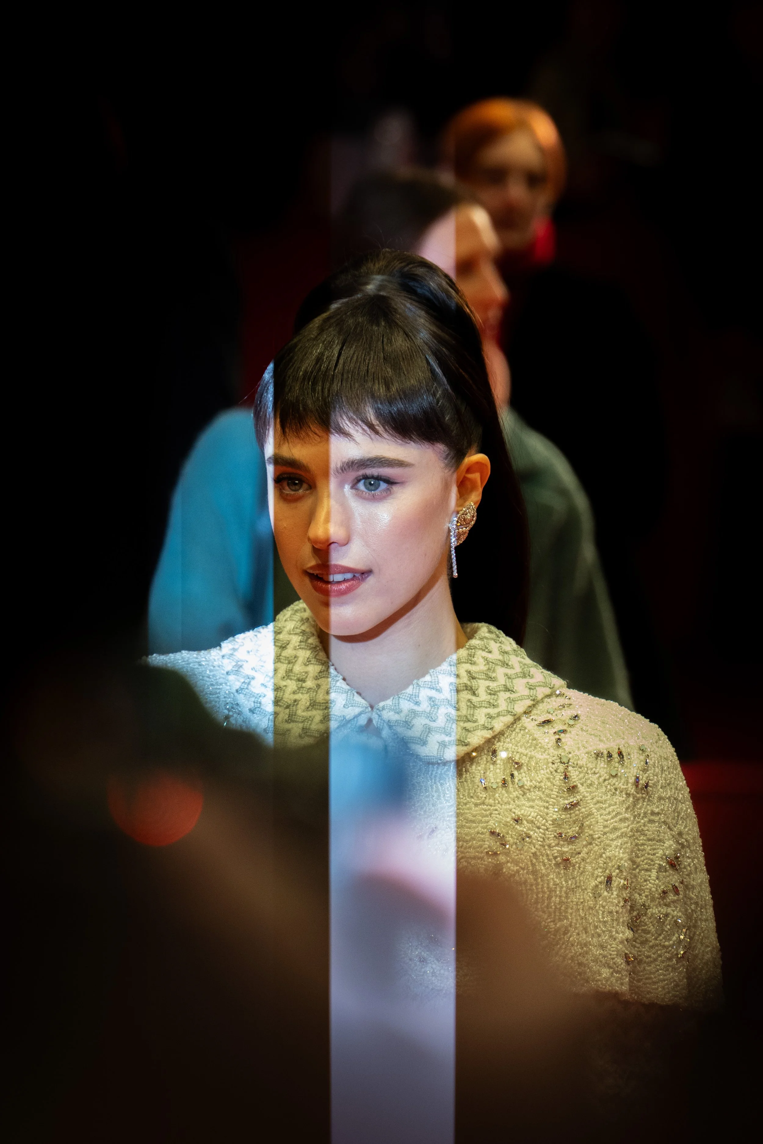 Eine Person mit dunklem Haar und schickem Outfit auf einem Event. Der Vordergrund ist verschwommen, und es gibt eine spiegelartige Reflexion über dem Gesicht.