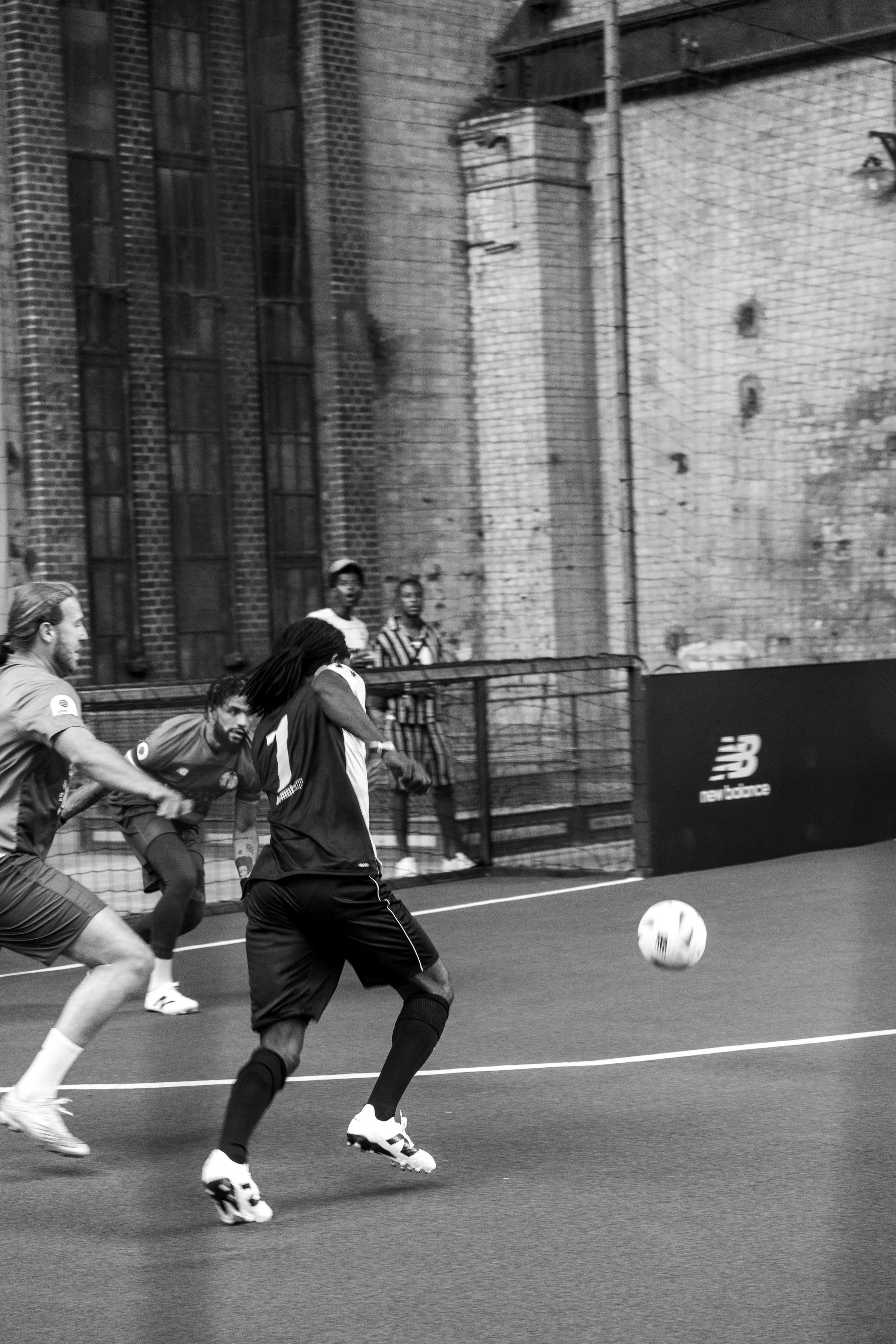 Schwarz-Weiß-Foto eines Fußballspiels in einer Halle mit drei Spielern im Vordergrund, einem Mann mit Rastazöpfen am Ball, während zwei andere versuchen, ihn zu verteidigen. Im Hintergrund sind Ziegelwände und Zuschauer zu sehen.
