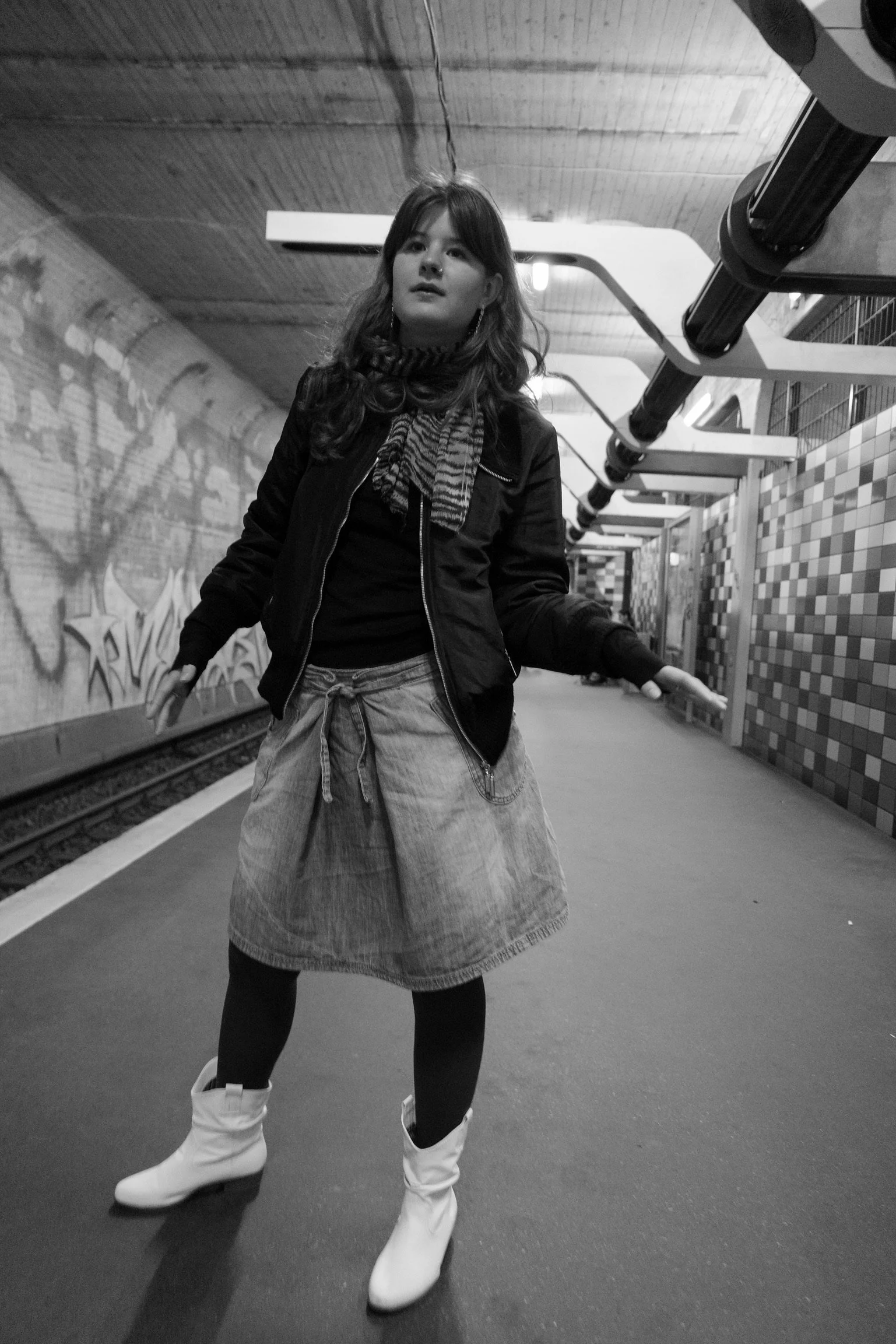 Eine Person steht in einer U-Bahn-Station. Sie trägt eine Jacke, einen karierten Schal, einen Jeansrock, dunkle Strumpfhosen und weiße Stiefel. Der Hintergrund zeigt Wandfliesen und Gleise.