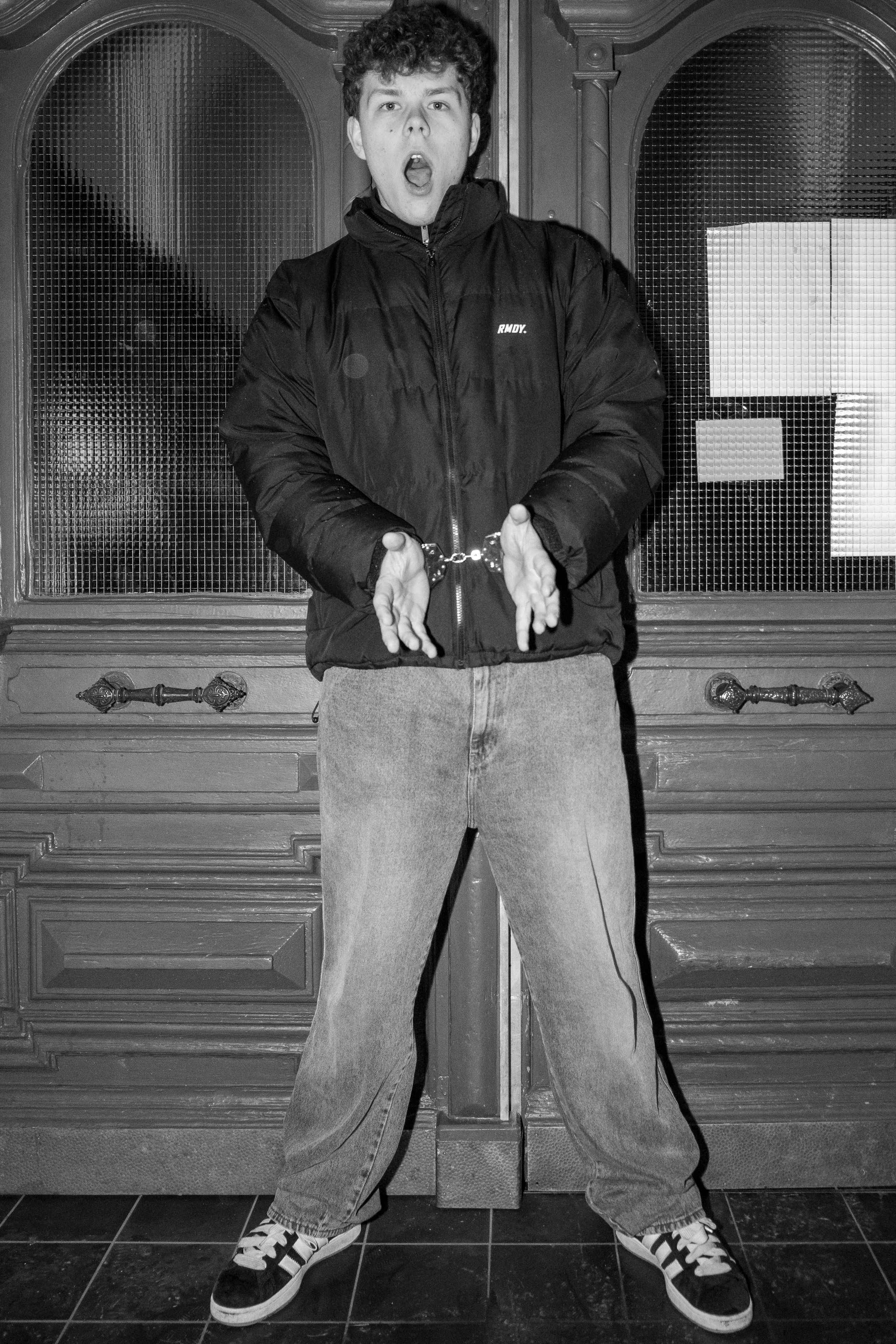 Eine Person in einer dicken Jacke und Jeans steht mit offenen Händen in Handschellen vor einer Holztür.