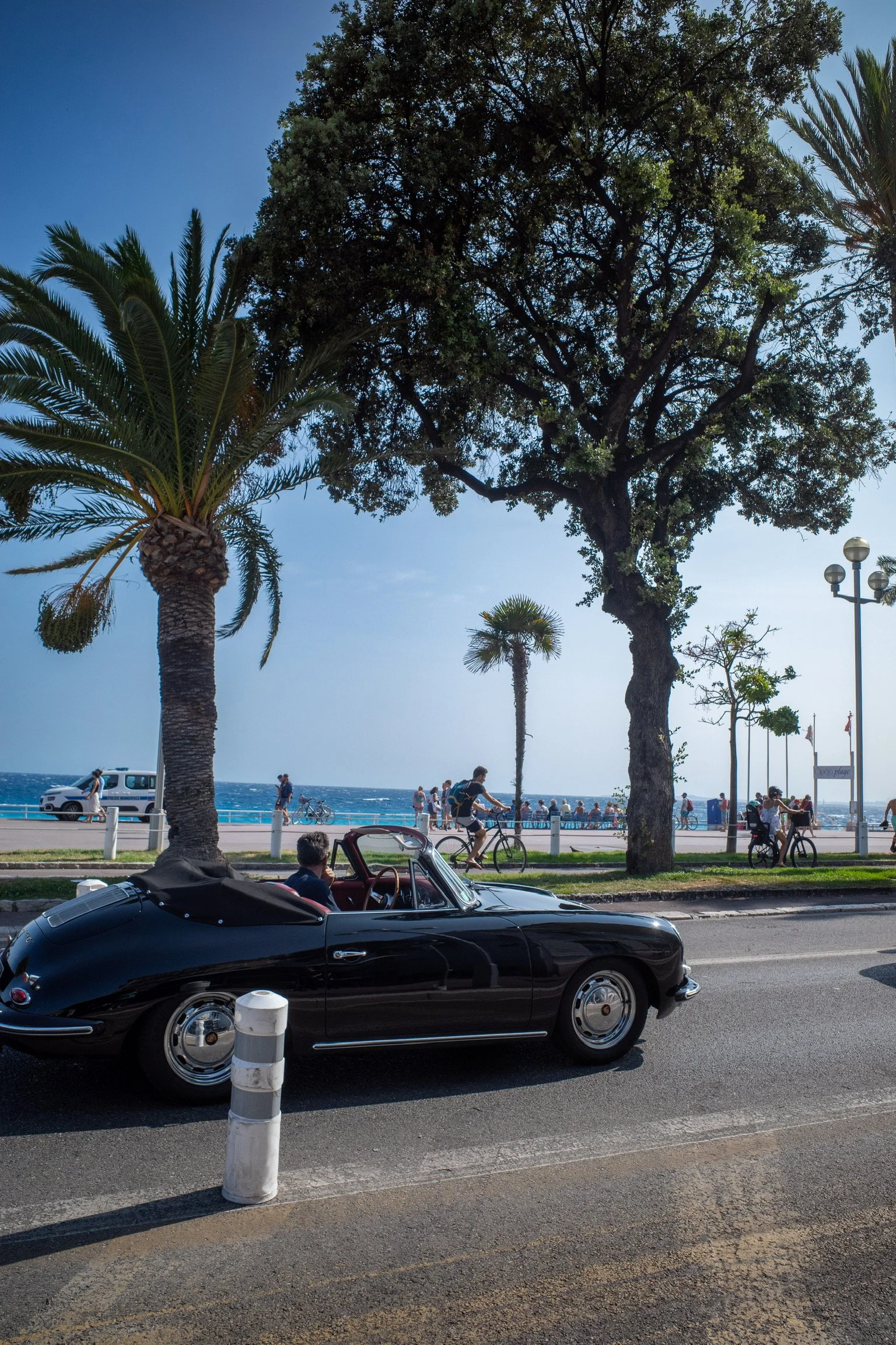 Schwarzes Cabriolet fährt auf einer Straße entlang einer Küstenpromenade mit Palmen, im Hintergrund Fahrradfahrer und Spaziergänger am Meer.