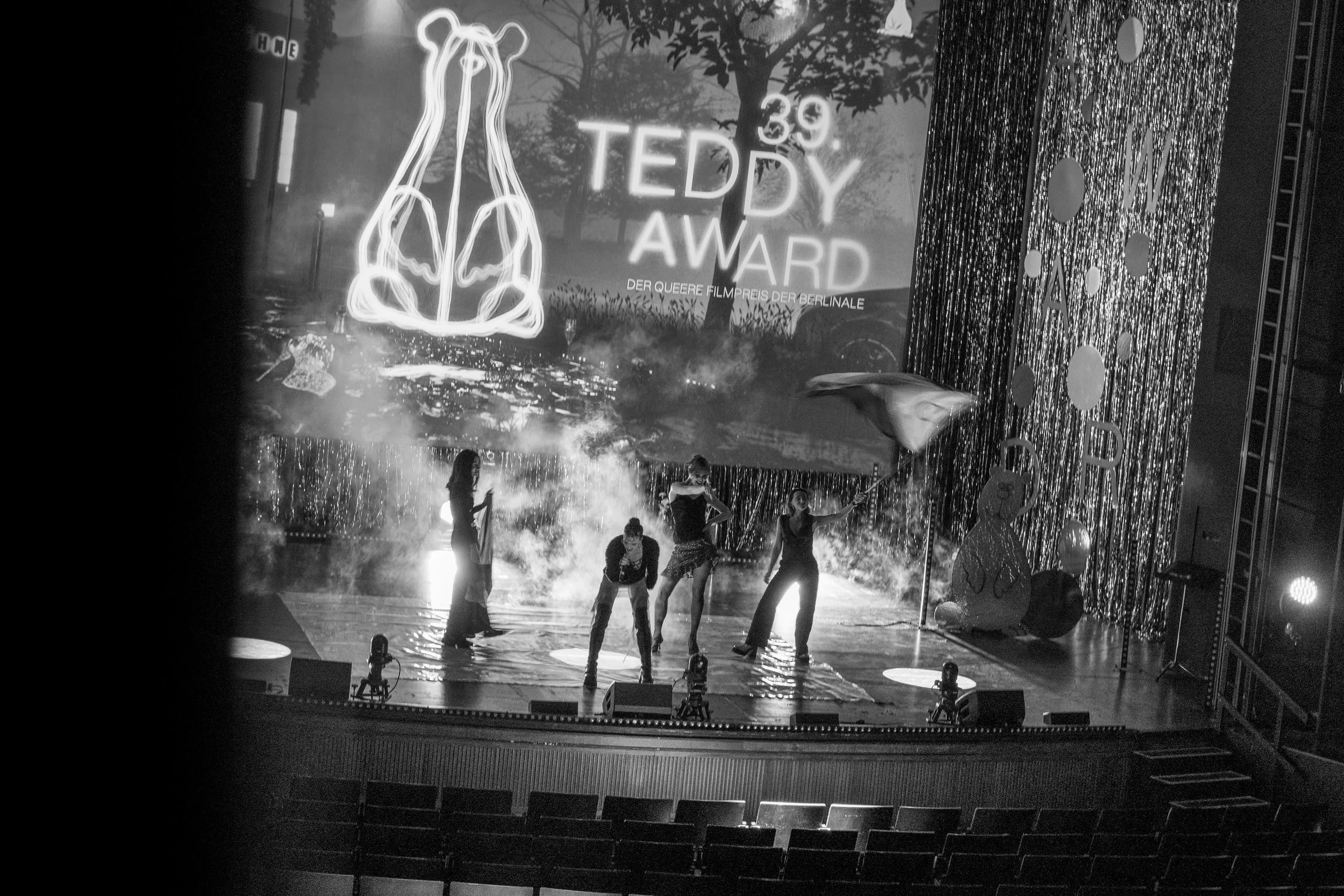 Schwarz-Weiß-Bild einer Bühne mit Personen, die vor einem großen Plakat mit der Aufschrift "39. Teddy Award" stehen, im Hintergrund bunte Dekorationen und Nebel.