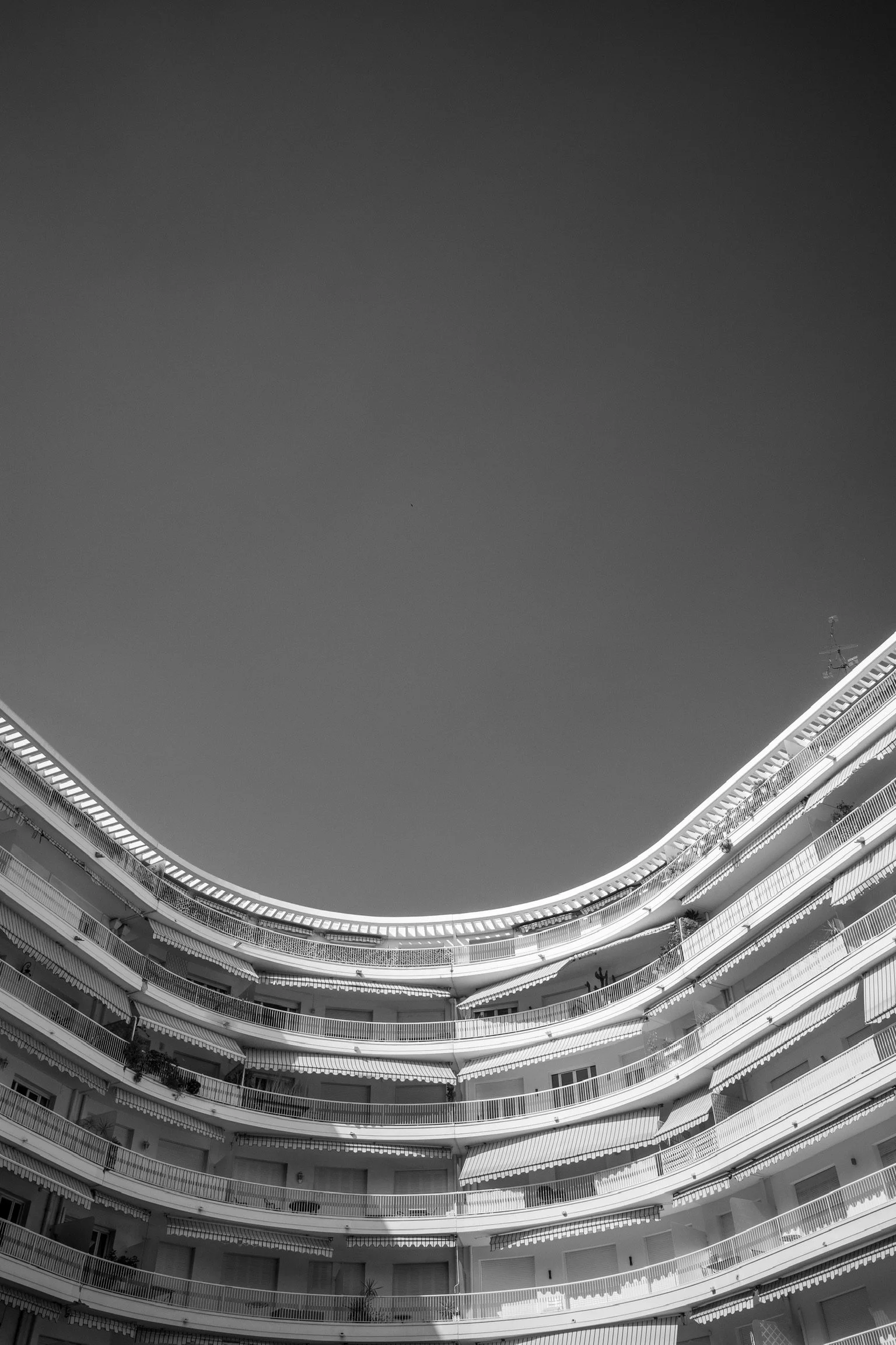 Schwarzweißaufnahme eines mehrstöckigen, runden Wohngebäudes mit Balkonumrandungen. Der Himmel ist klar und dunkel, Gebäude hat gebogene Architektur.