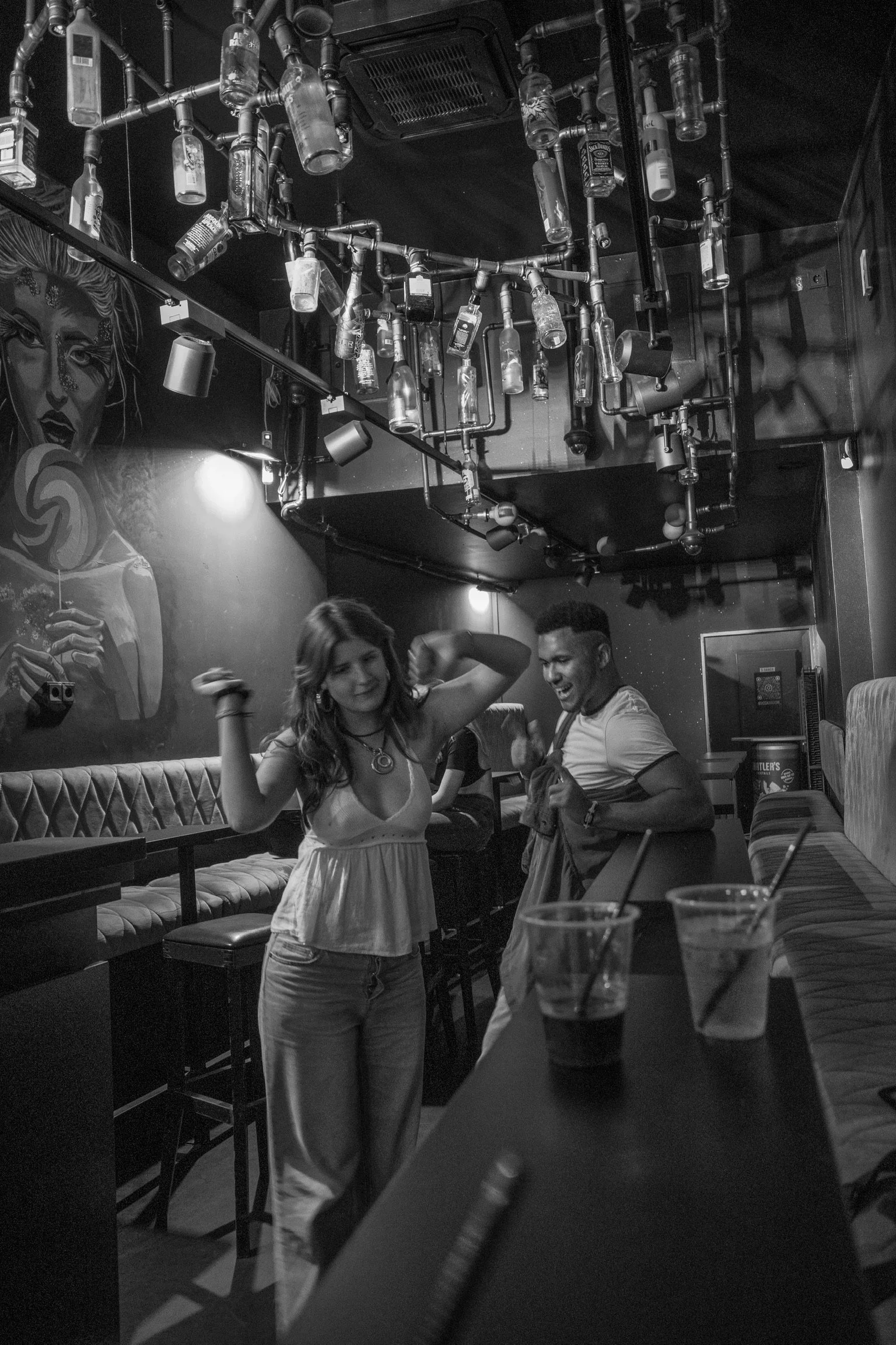 Schwarz-Weiß-Foto von einer Bar mit tanzenden Personen, Flaschen als Dekoration an der Decke und modernen Kunstwerken an den Wänden.