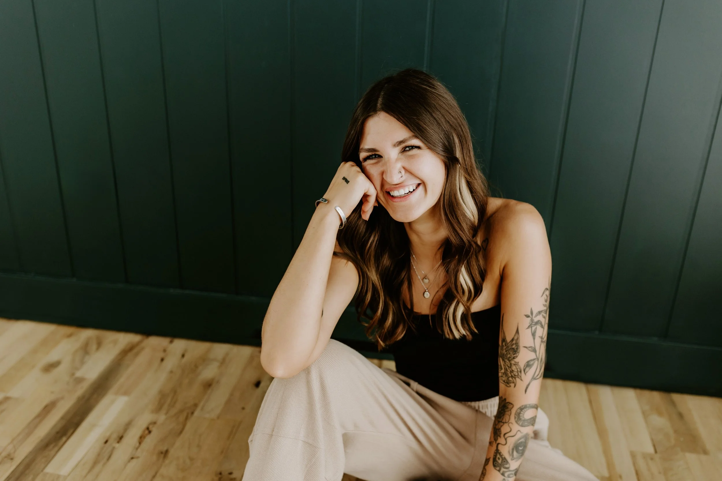 A person with long brown hair, wearing a brown long-sleeve top and light jeans, sitting on a chair in a bright room. They are smiling and touching their hair.