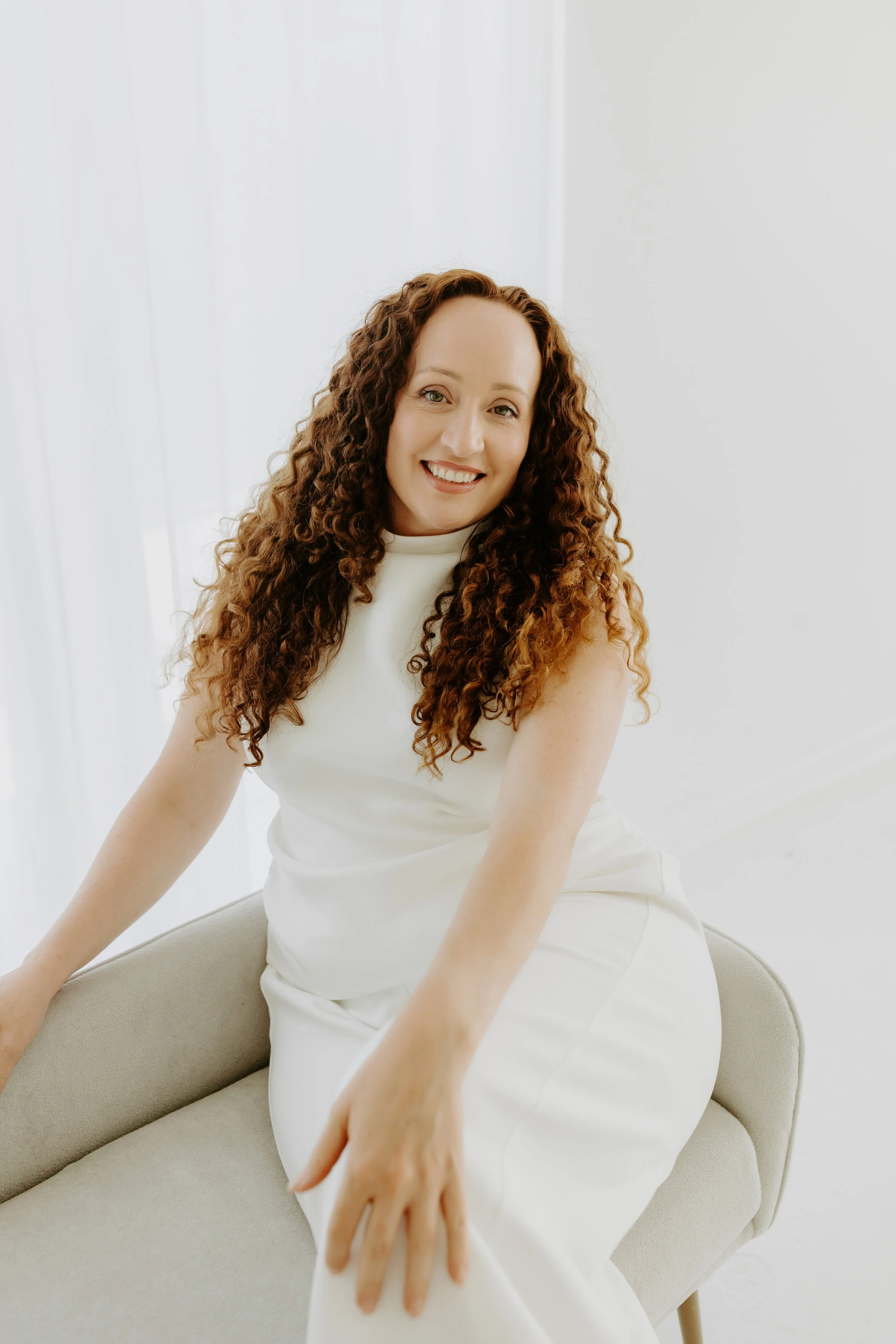 A woman with curly brown hair standing indoors on a wooden floor next to a wooden stool, wearing a white top and beige wide-leg pants.