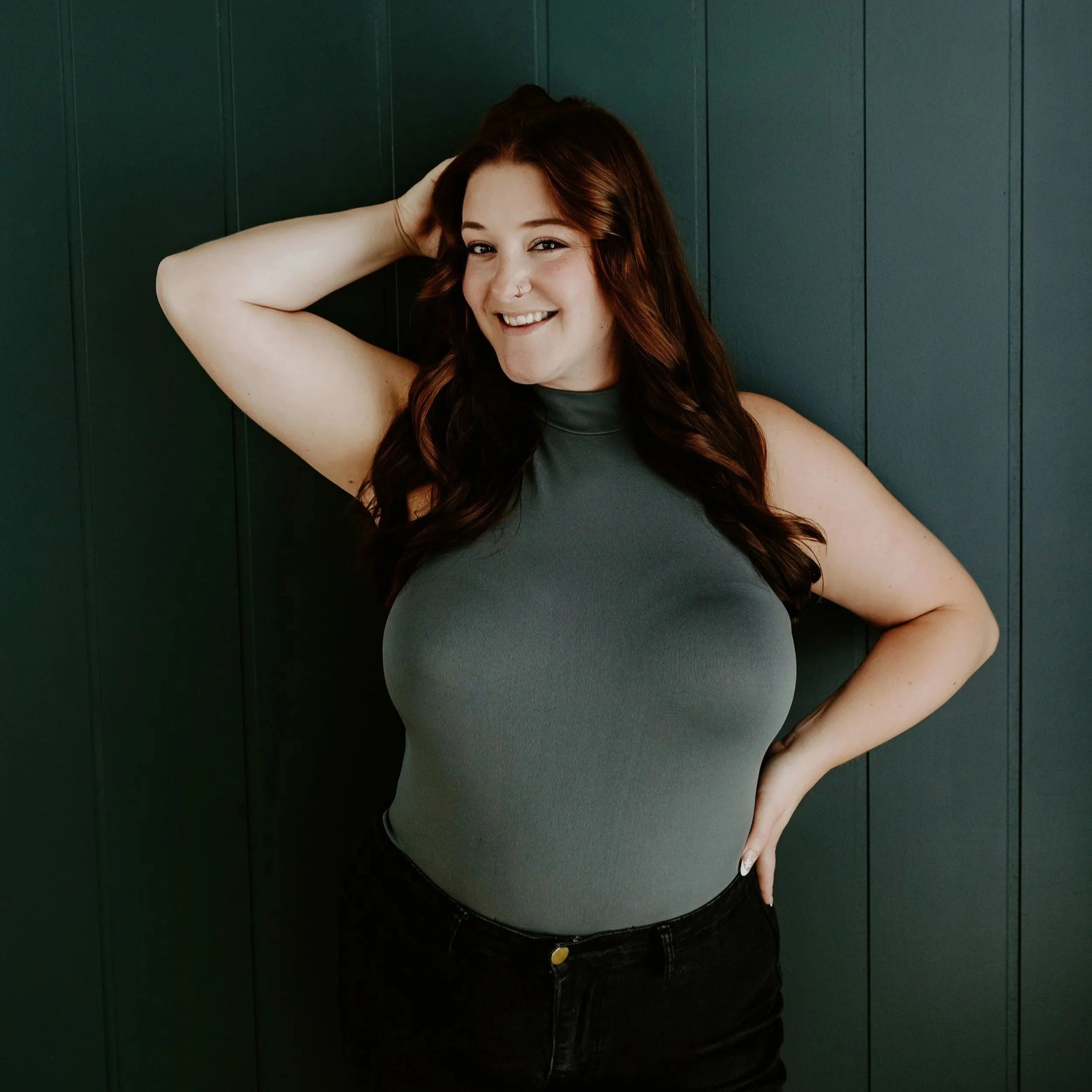 A smiling woman with wavy red hair, wearing a beige sleeveless top, standing against a plain white background.