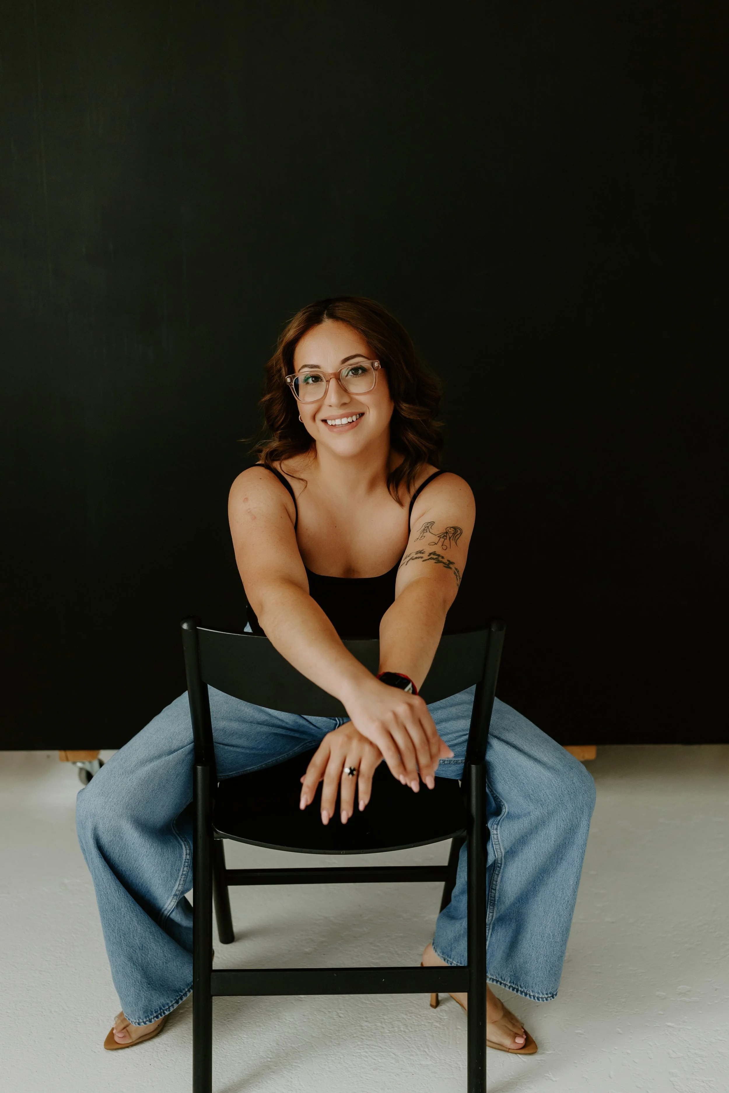 Woman smiling seated in a chair, wearing a beige blazer and jeans, with long brown hair and natural makeup.