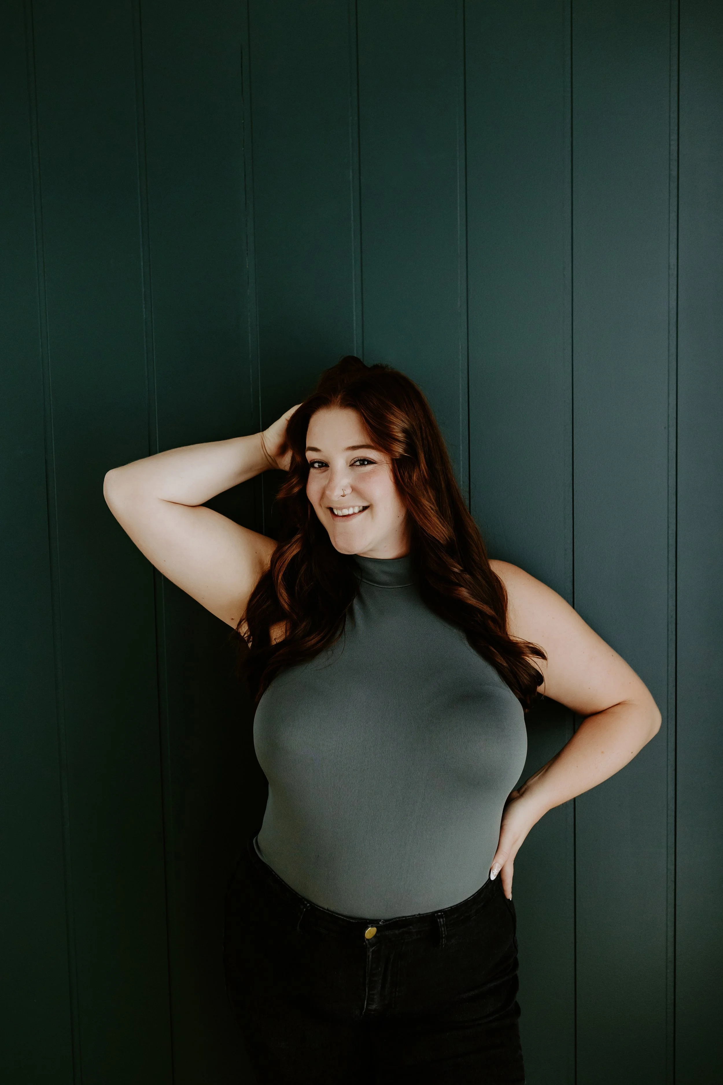 A smiling woman with wavy red hair, wearing a beige sleeveless top, standing against a plain white background.