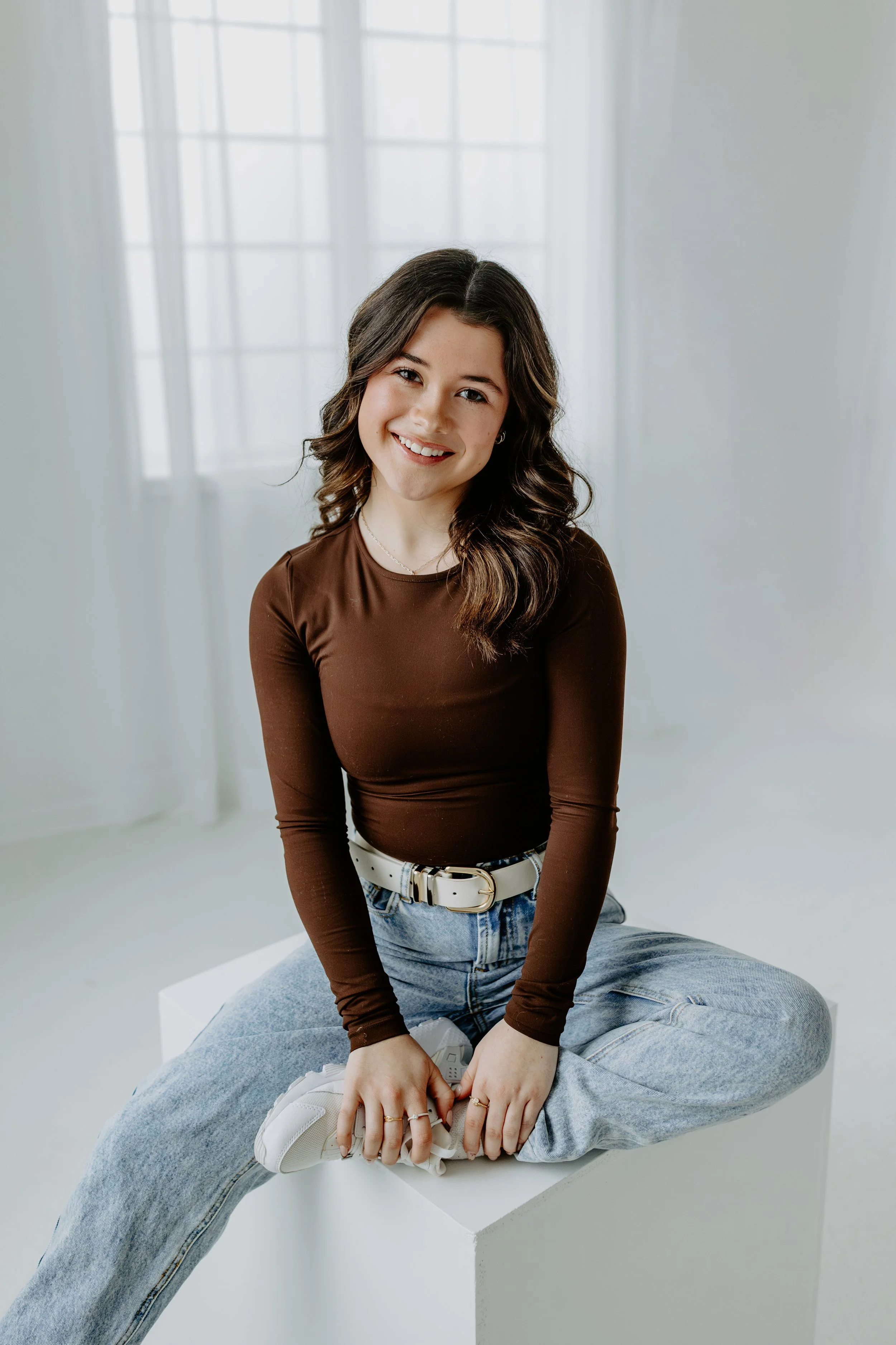 Young woman with dark wavy hair, light skin, wearing clear glasses, a black t-shirt, and a gold necklace with a letter D pendant, smiling in front of a brick wall.