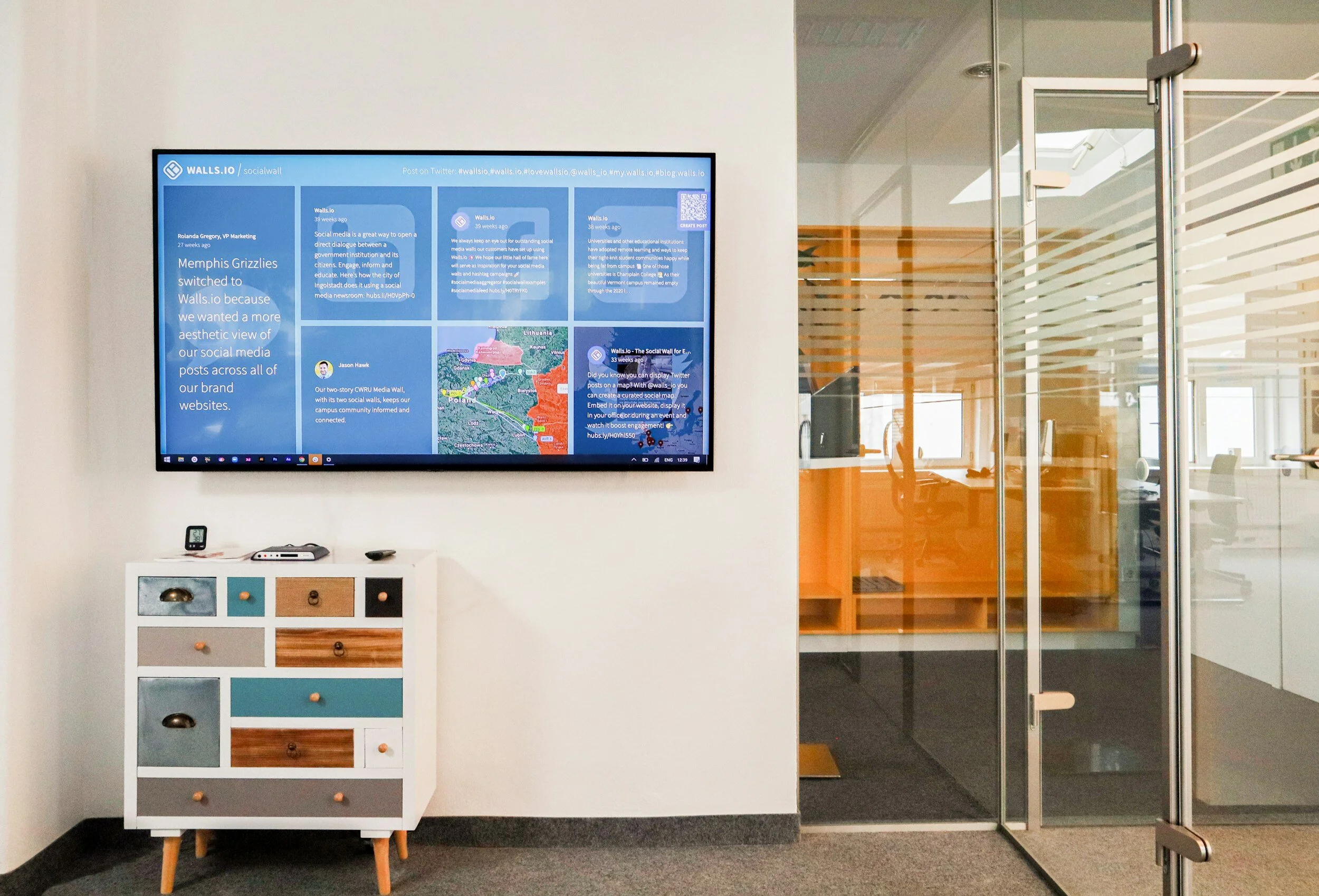 Office lobby with a large digital screen displaying social media posts, a colorful drawer unit beneath the screen, and a glass door leading into a modern office space with orange accents.