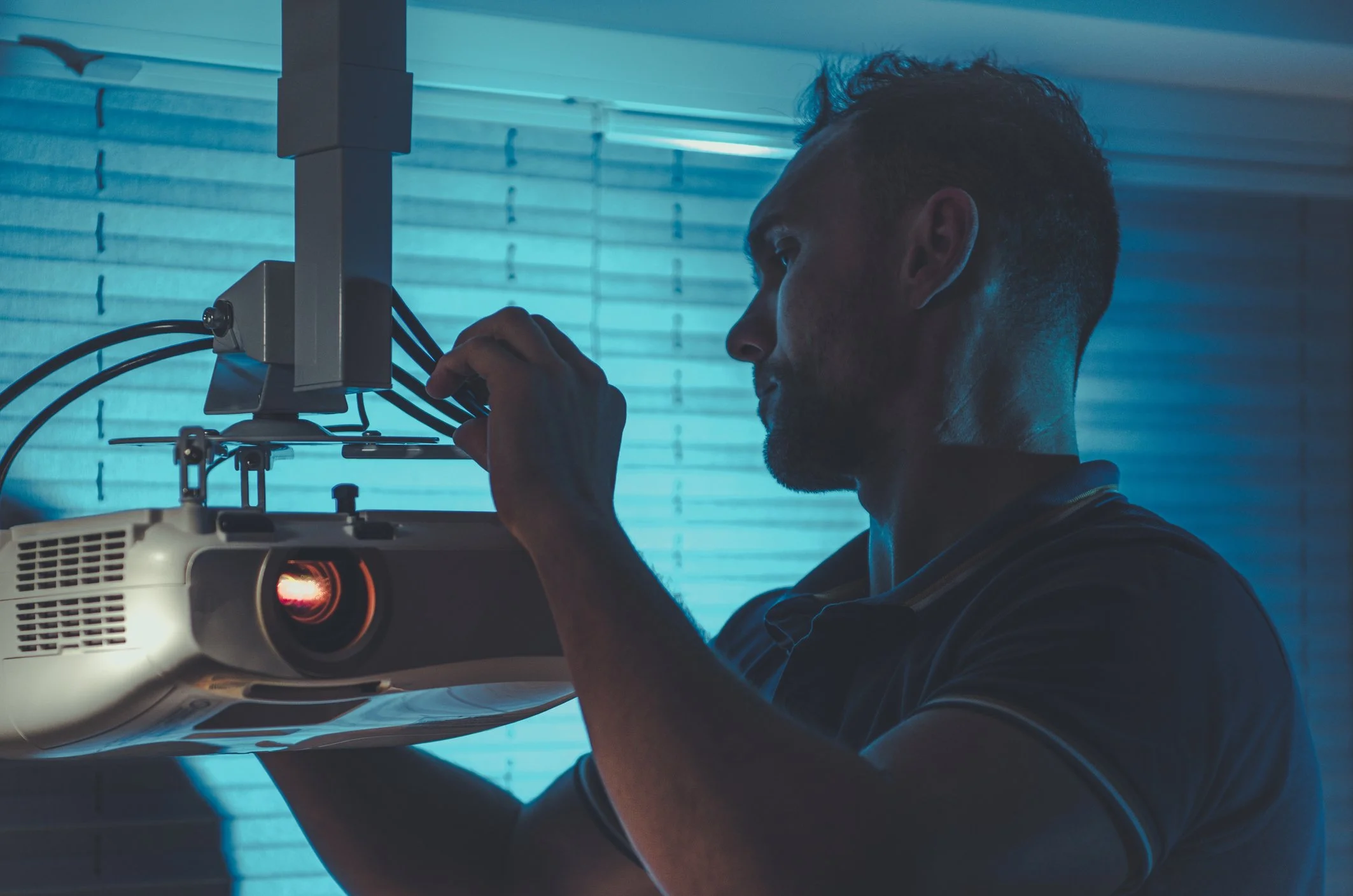 A man adjusting a projector in a dimly lit room with blue blinds.
