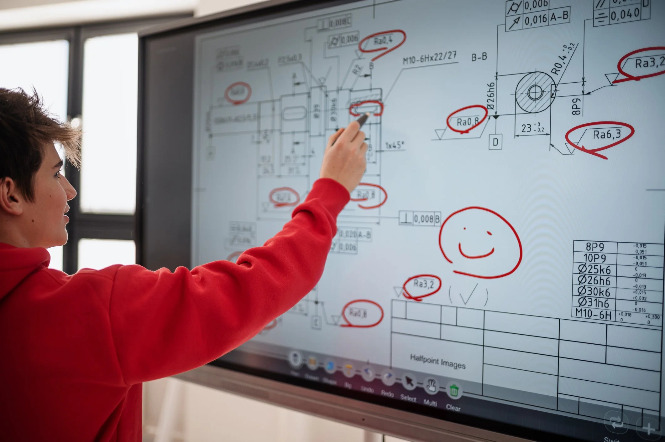 A young man in a red hoodie points at a technical engineering drawing on a large display, with red circles highlighting certain areas and a red smiley face drawn on the screen.