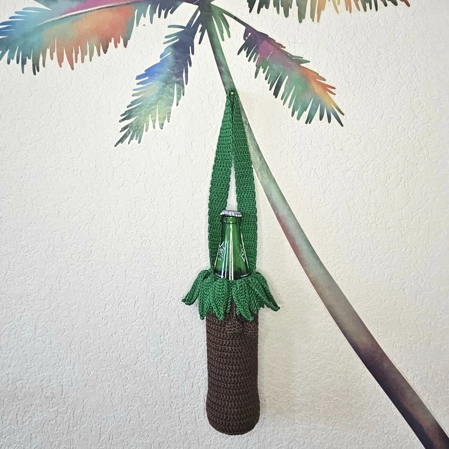 A crocheted hanging bag resembling a tropical drink, with a green bottle inside, hanging from a crocheted palm tree wall decoration.