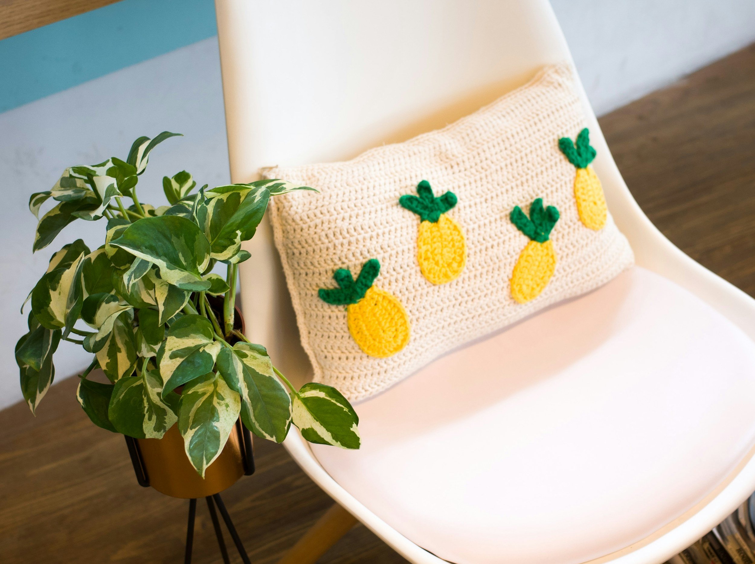 A white chair with a crochet pillow decorated with yellow pineapples with green leaves, next to a potted plant with variegated green and white leaves.