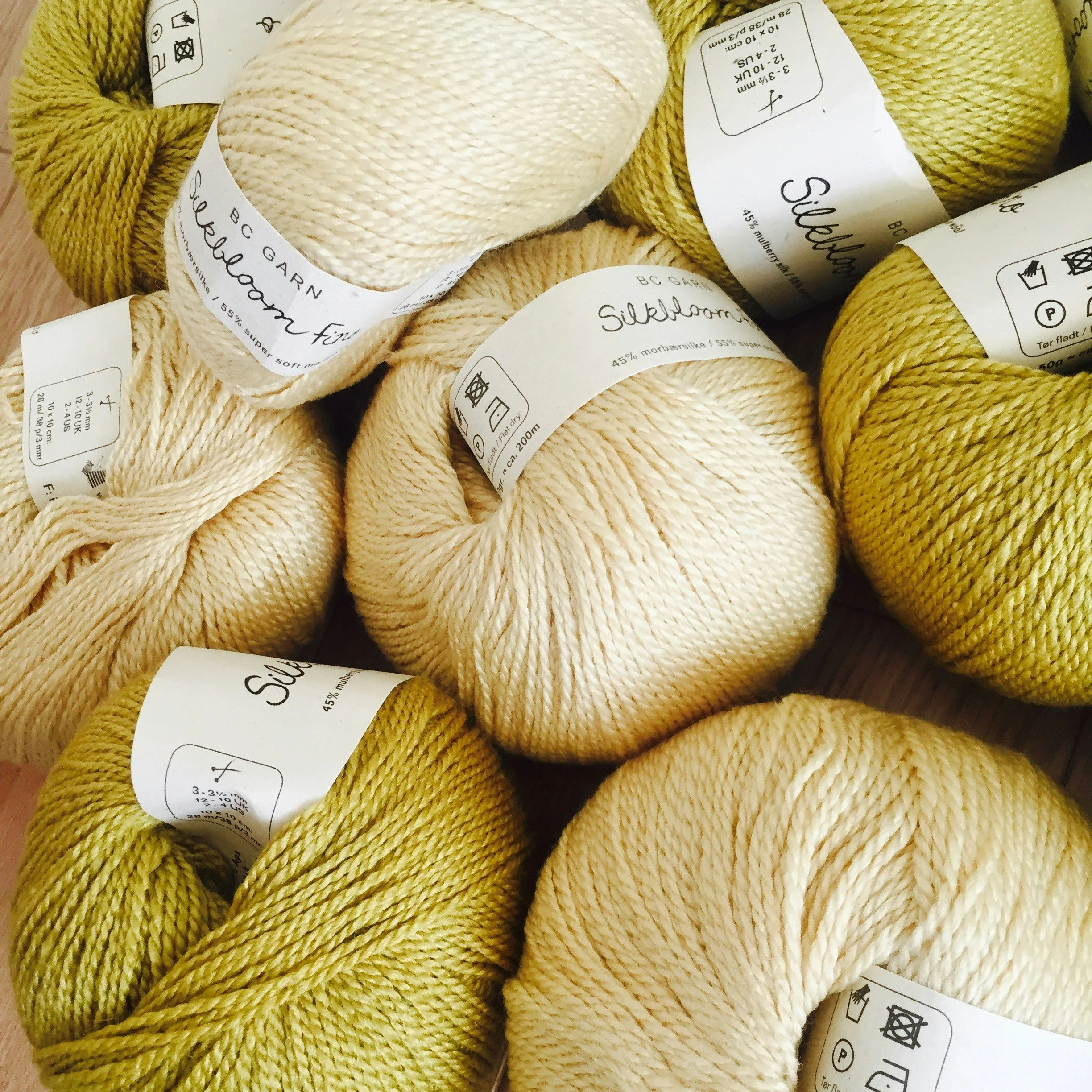 Various skeins of cream and yellow colored yarn piled together, labeled with brand and fiber content.