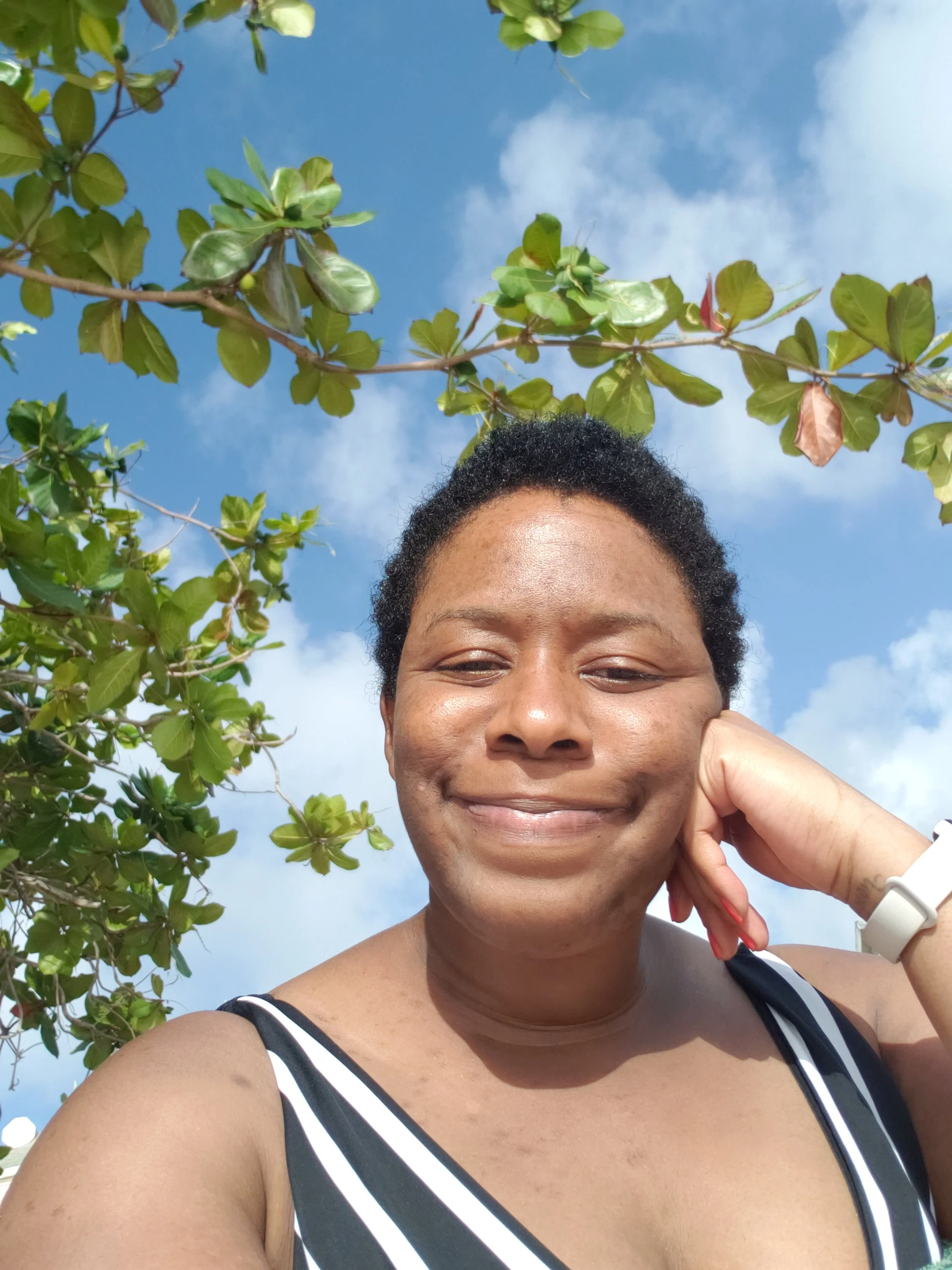 A woman smiling outdoors against a blue sky with some clouds, resting her head on her hand, with green leafy branches above her.
