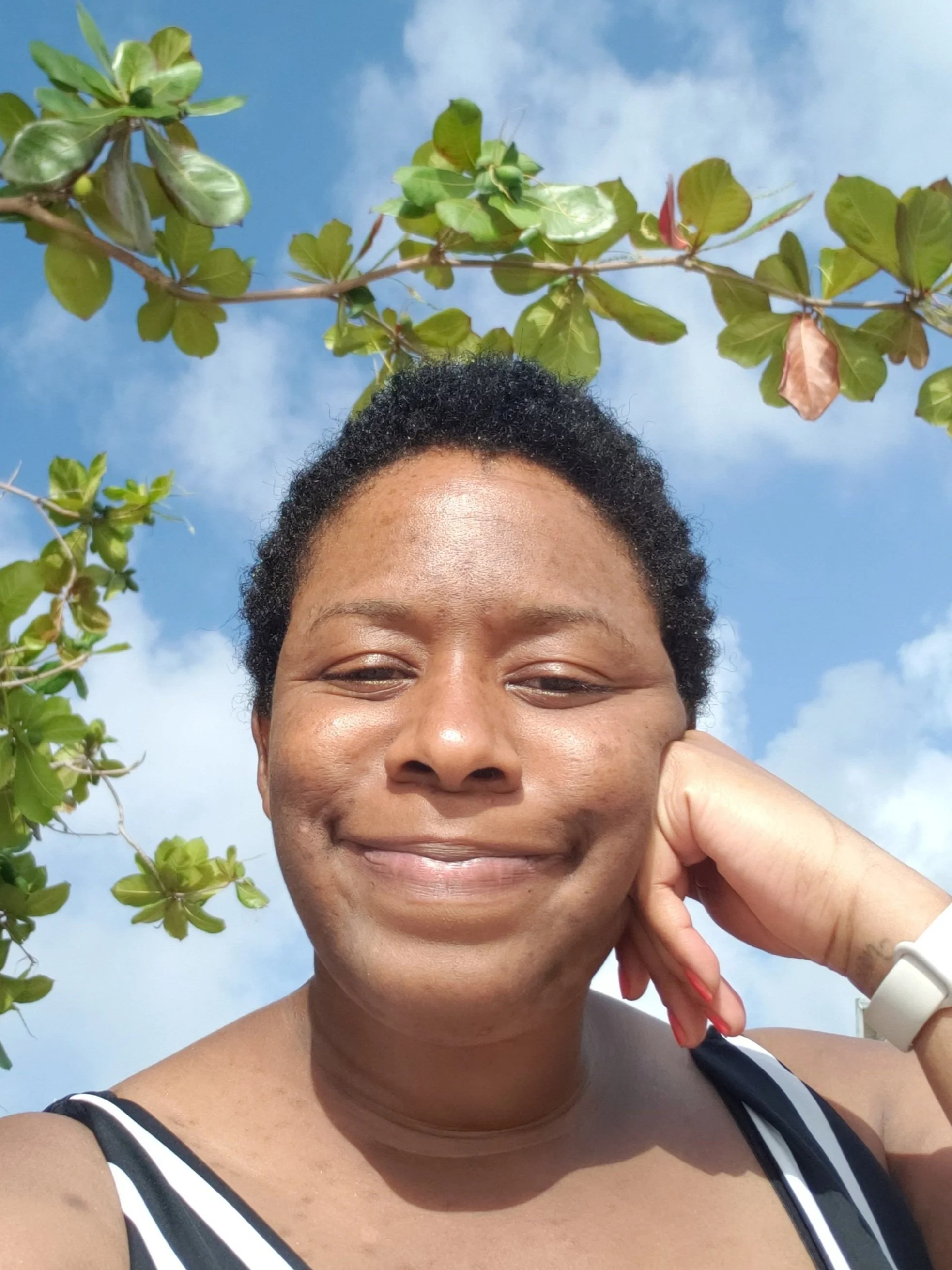 A woman smiling outdoors, resting her face on her hand, with a bright blue sky and green leafy tree branches in the background.