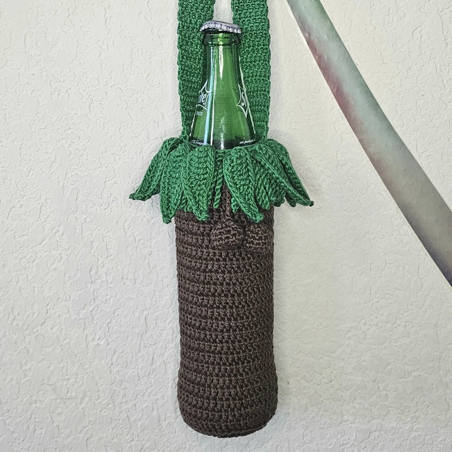 Crocheted object resembling a plant with brown stem and green leaves, holding a glass bottle.