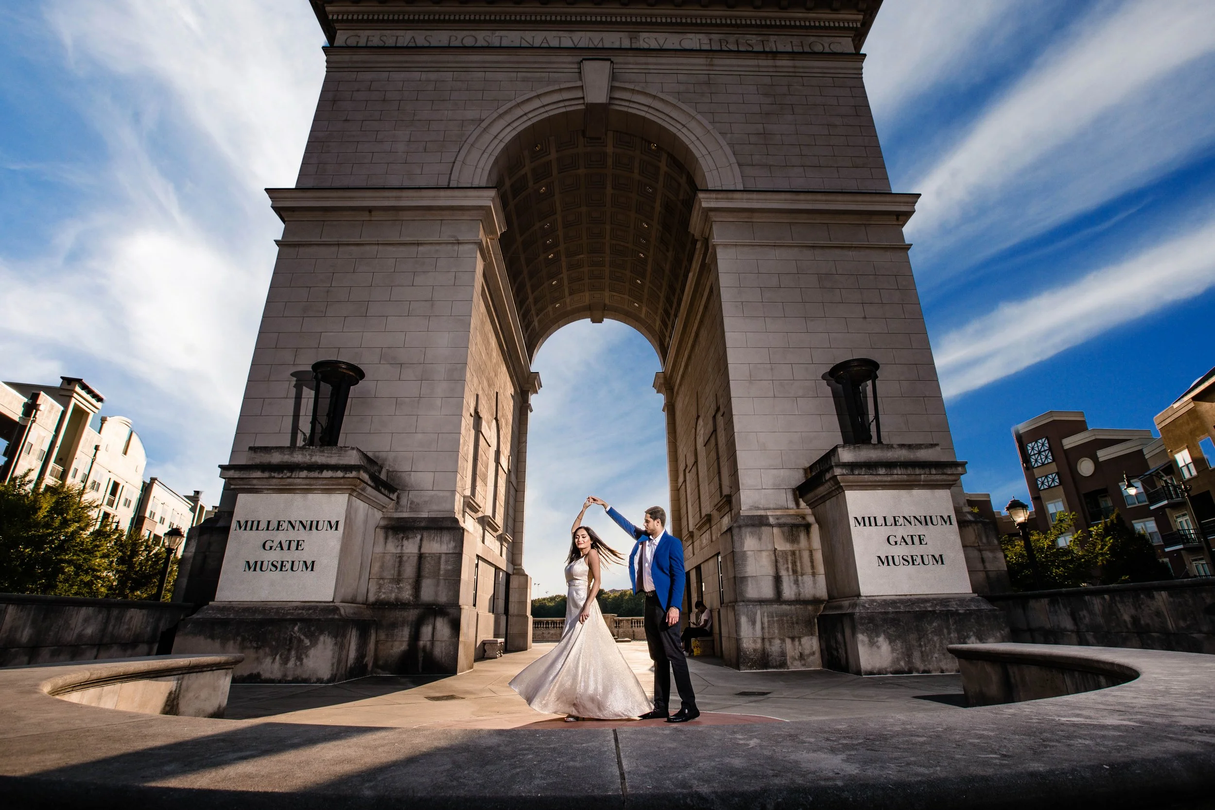 A bride and groom dancing under a large stone archway at the Millennium Gate Museum on a sunny day.