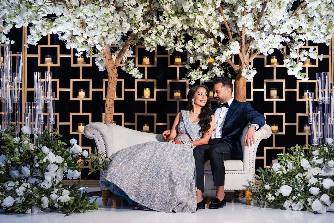 A couple in formal attire sitting on a white sofa surrounded by white flowers and greenery, with decorative candles and trees behind them, at an elegant event or wedding.