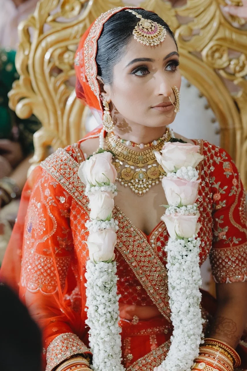 A bride in traditional Indian attire, wearing intricate gold jewelry and red embroidered clothing, adorned with a floral garland of white roses, during a wedding ceremony.