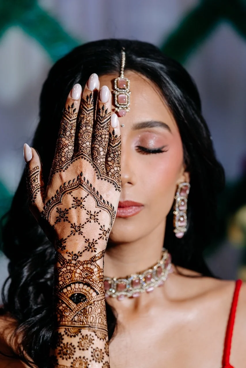 A woman with dark hair and makeup, dressed in traditional jewelry, covering one eye with her hand decorated with intricate henna designs.
