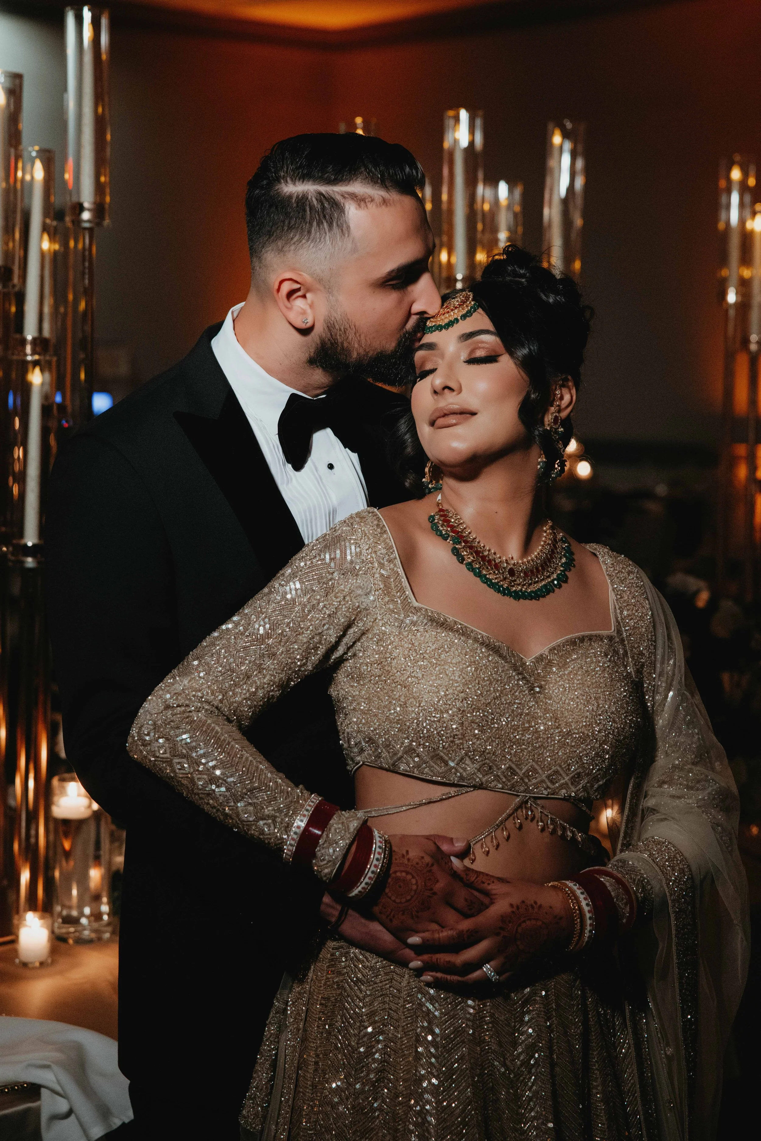 A couple at a wedding, the man in a black tuxedo and the woman in a gold traditional Indian dress, the man kissing the woman's forehead.
