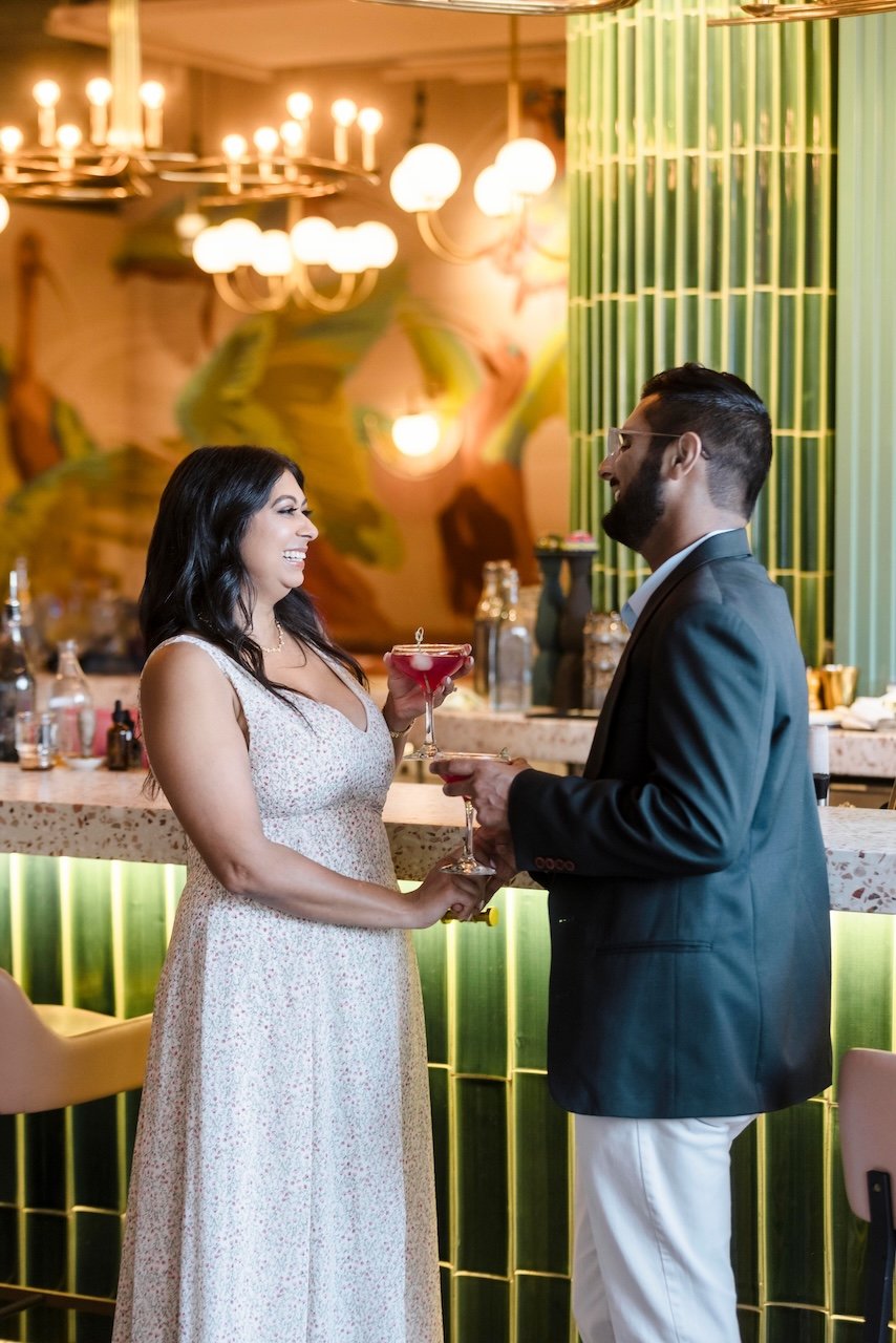 A couple at a bar, holding hands and smiling at each other, with drinks in hands. The woman wears a light-colored dress, the man wears a dark blazer and white pants. The bar has a green and yellow tile design and a well-lit background with chandelier
