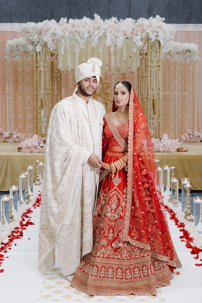 Indian bride and groom in traditional wedding attire standing on decorated aisle with candles and flower petals.