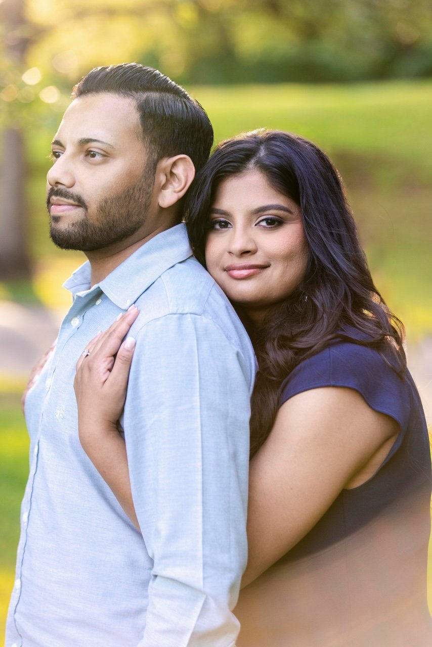 A man and woman standing close together outdoors, with the woman resting her head on the man's back, both smiling softly.