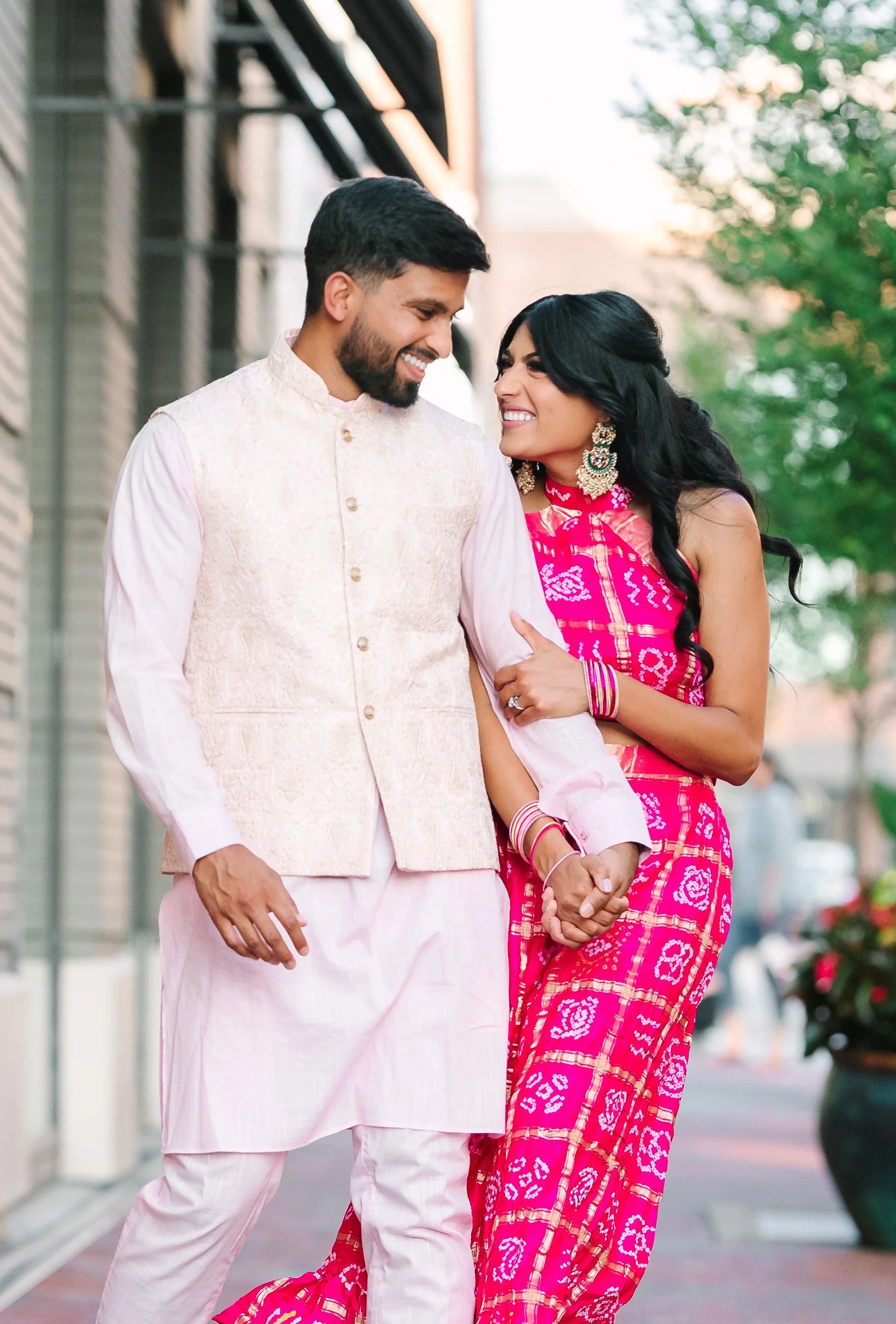 A couple dressed in traditional Indian attire, holding hands and smiling at each other outdoors on a city sidewalk.
