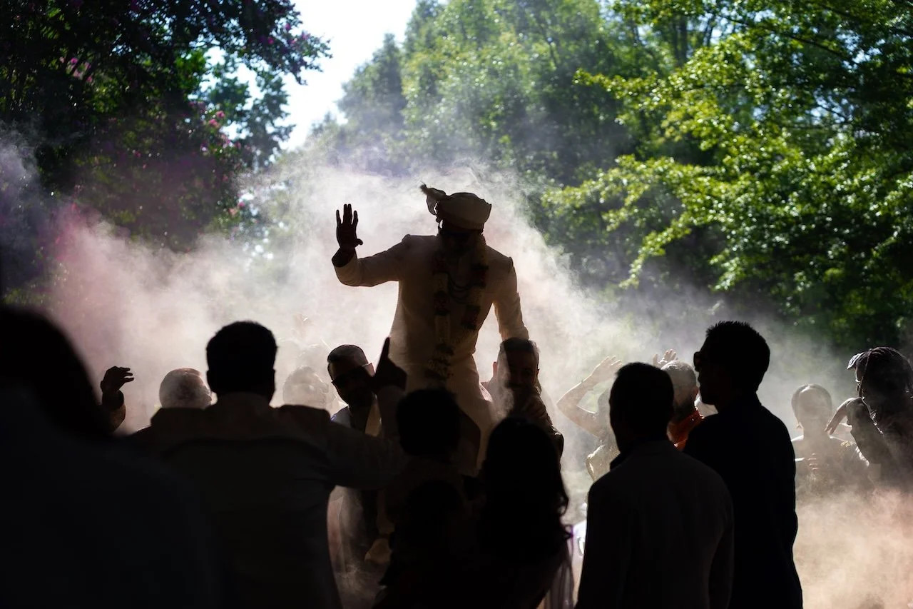 A silhouette of a man dressed in traditional attire riding on shoulders of others amidst a cloud of dust or powder, surrounded by a crowd of people, in an outdoor setting with lush green trees.