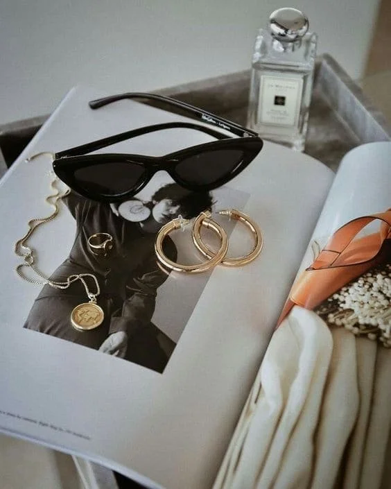 A pair of black sunglasses, gold hoop earrings, a gold necklace with a pendant, and a ring are arranged on a black and white photo of a woman. A bottle of perfume and a beige and orange patterned scarf are also on the table.
