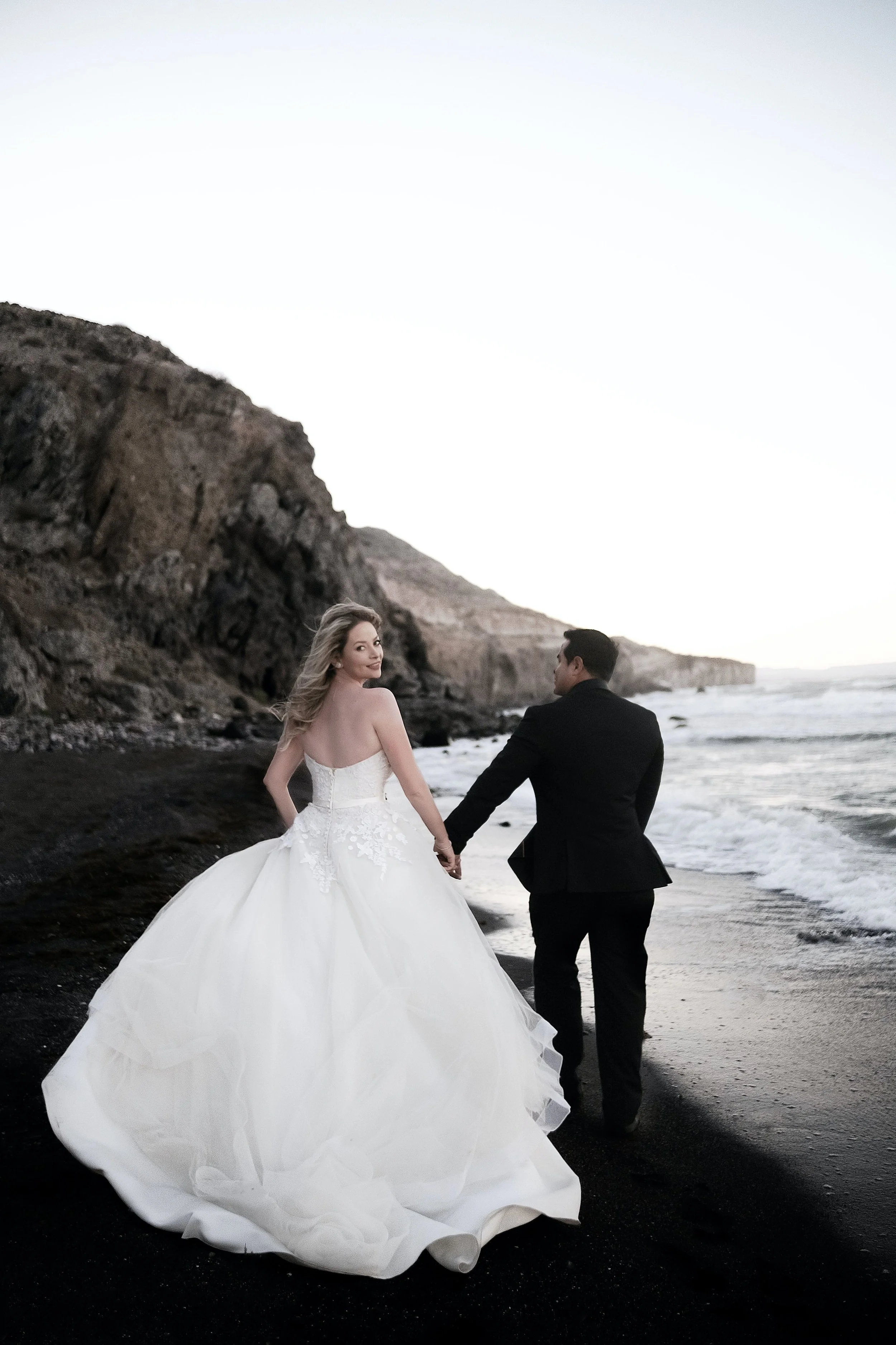 A bride and groom walking hand in hand on a beach at sunset or sunrise, with rocky cliffs in the background.