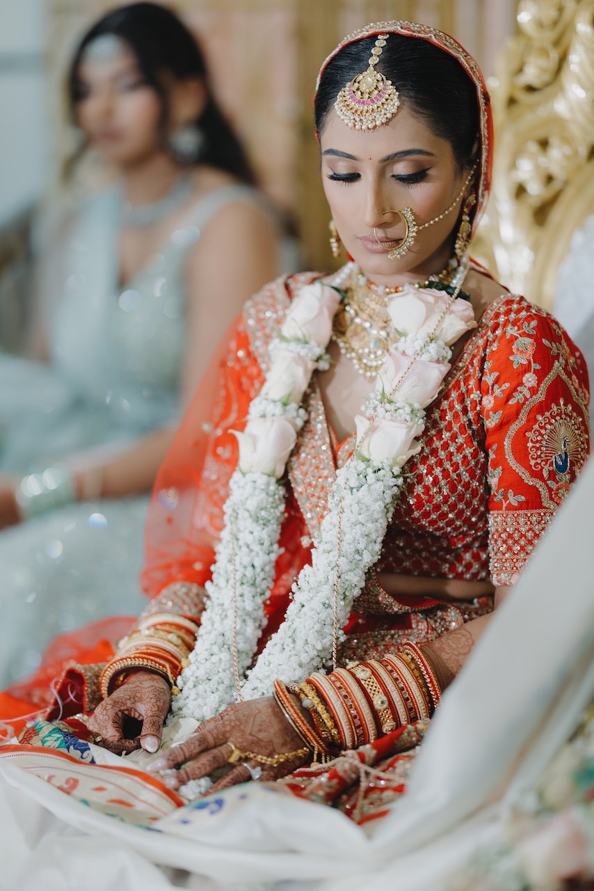 A bride dressed in traditional Indian wedding attire, wearing red and gold embroidered clothing, elaborate jewelry including a maang tikka, nose ring, and bangles, and holding a flower garland, during her wedding ceremony.