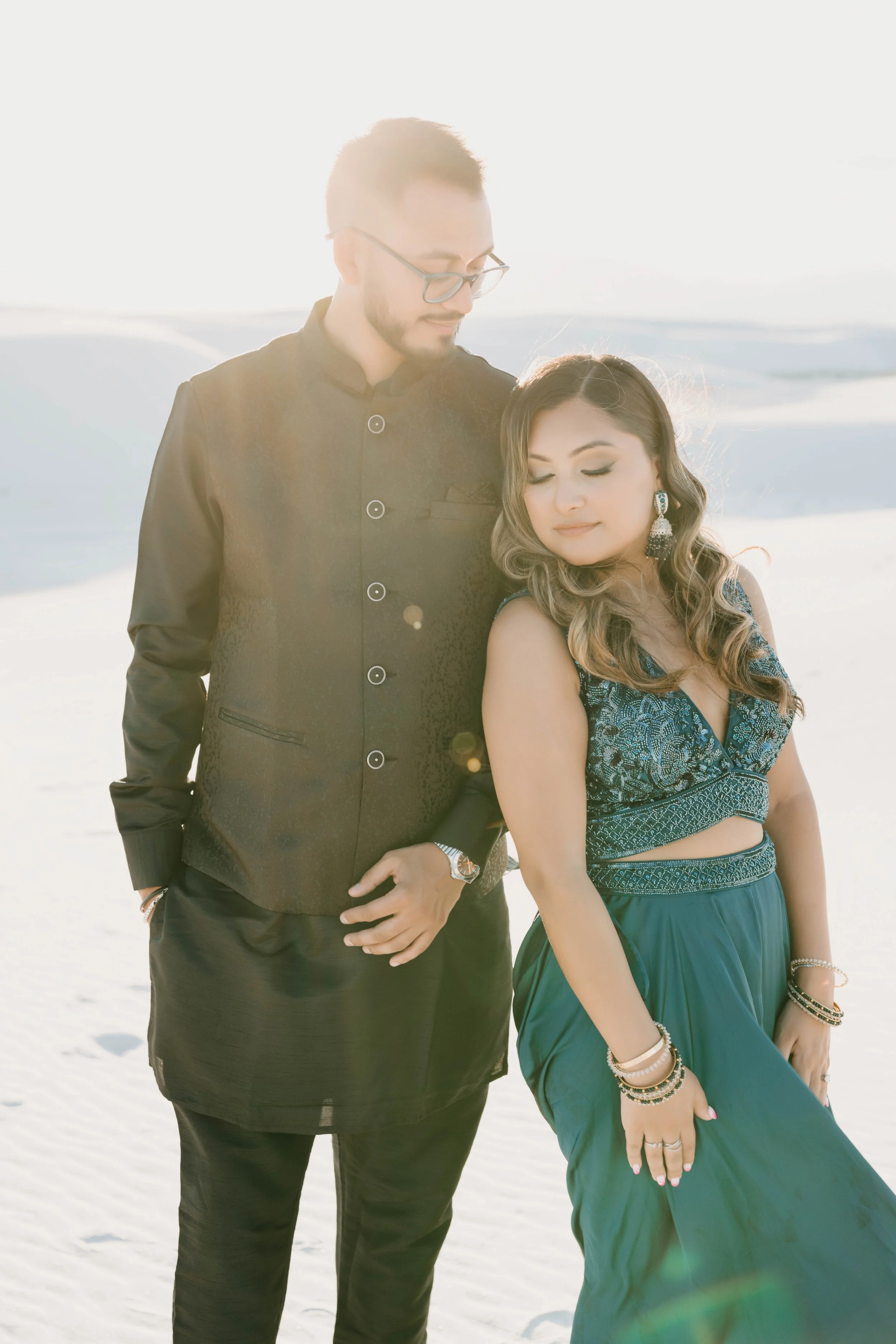 A man and woman dressed in traditional Indian attire standing close together on a bright sandy beach, with the man looking down at the woman and the woman with closed eyes, surrounded by sunlight.