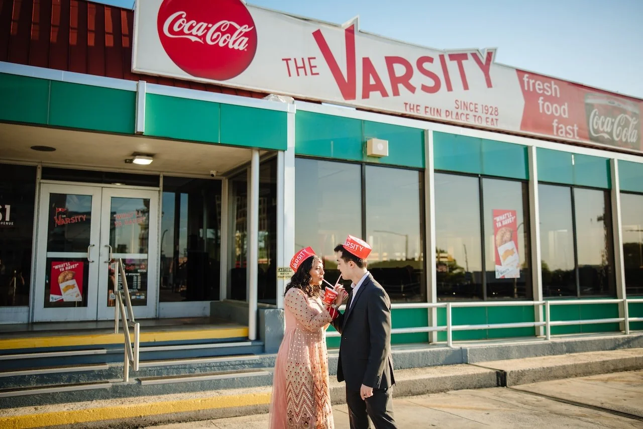 A young couple dressed nicely, wearing Varsity paper hats, sharing a drink outside a fast food restaurant named The Varsity. The restaurant has a large Coca-Cola sign and promotional posters on the windows.