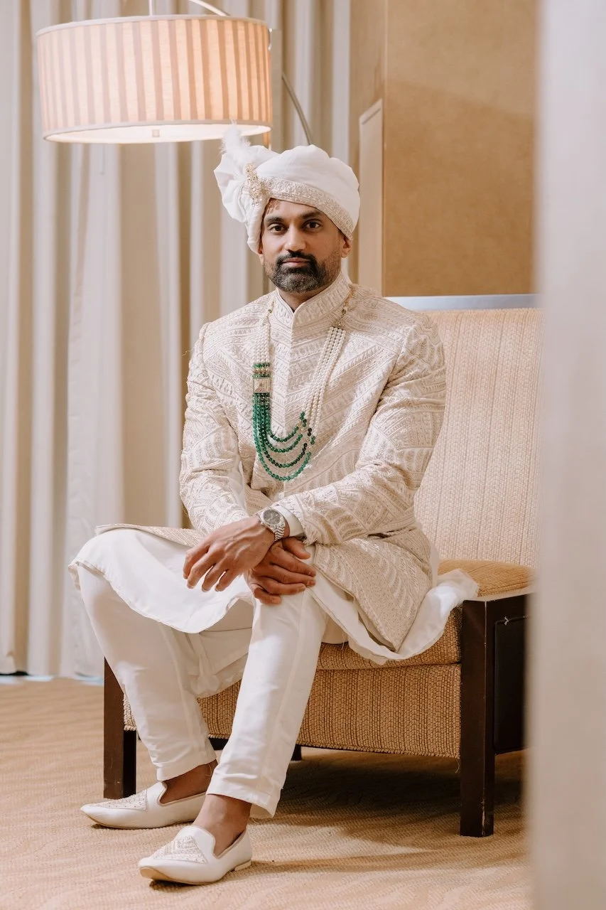 A man dressed in traditional Indian wedding attire, sitting on a chair in a hotel room. He is wearing a cream-colored sherwani with intricate embroidery, a matching turban with a feather accessory, green beaded necklaces, a wristwatch, and white shoe