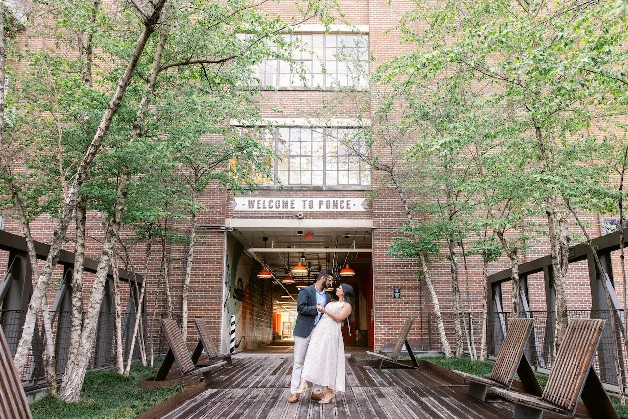 A couple dancing on a wooden walkway outside a brick building with green trees on either side. The building has a sign that reads 'Welcome to Ponce'.
