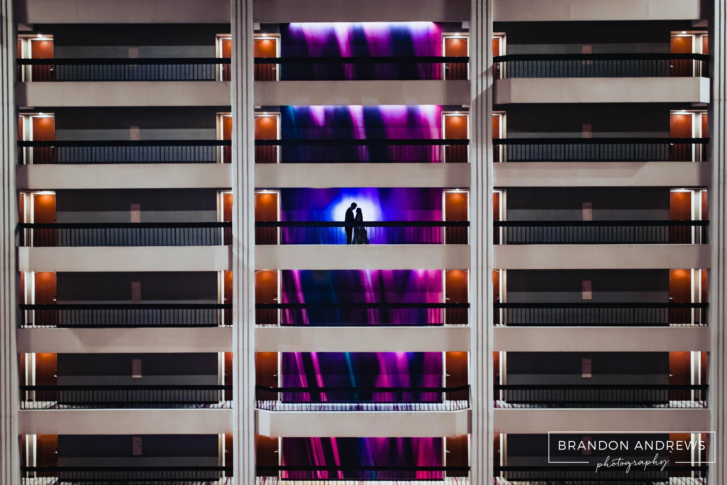 Silhouetted couple standing close on balcony of a hotel, with colorful purple and blue neon lights in the background, multiple hotel rooms with closed doors and balconies visible throughout the building.