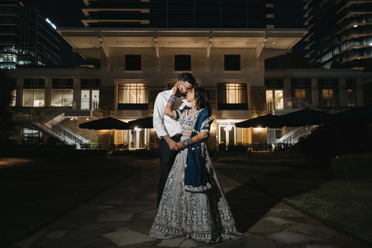 A couple standing close together during nighttime in front of a lit building, with the man wearing a white shirt and the woman in traditional Indian attire.