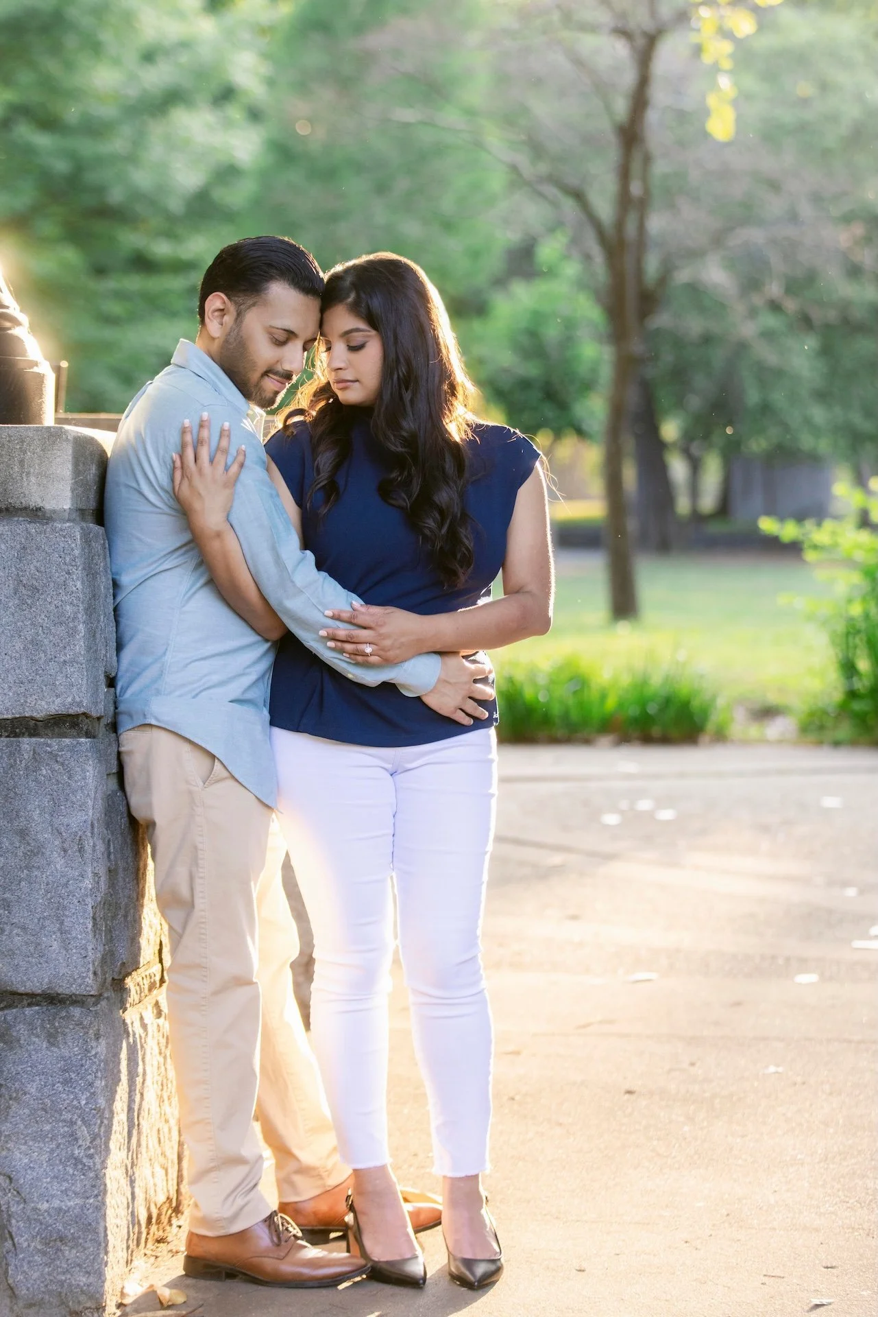 A young couple stands close together outdoors in a park during sunset, sharing an intimate moment with their foreheads touching, eyes closed, and arms around each other.