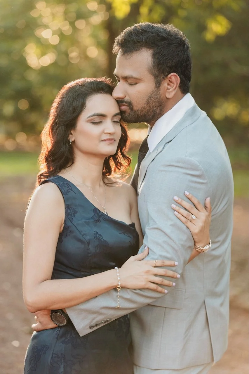 A romantic couple embracing outdoors during sunset, with the man gently holding the woman and their foreheads touching, eyes closed, surrounded by blurred green and yellow trees.