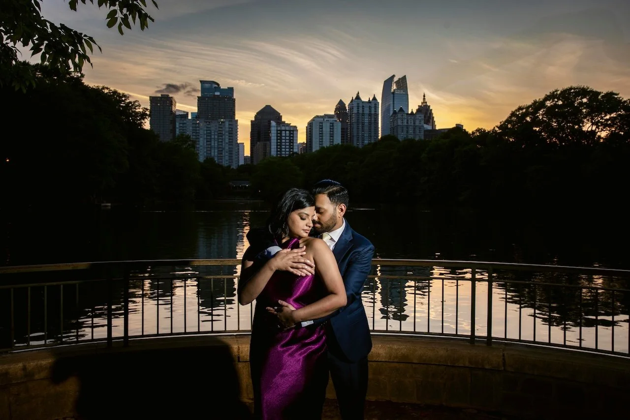 A couple embraces near a lake at sunset with a city skyline in the background.