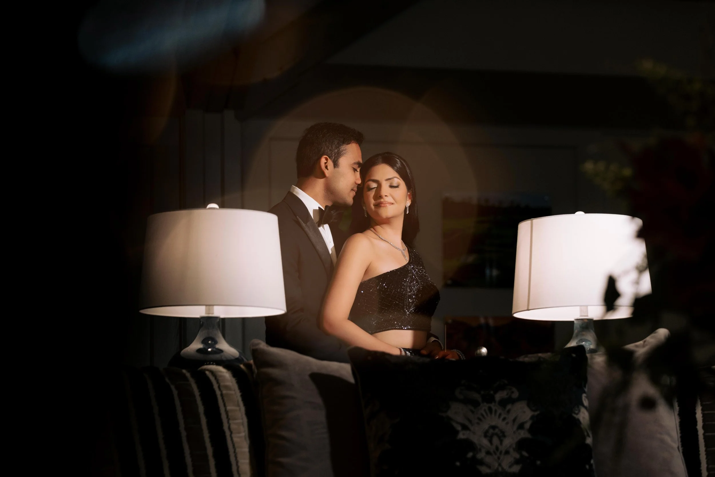 A young man and woman dressed in formal attire are sitting close together in a dimly lit room, with a mirror reflecting their intimate moment between two lamps.