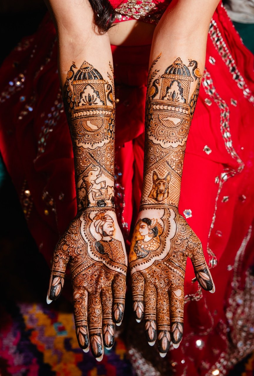 Intricate henna tattoo designs on woman's hands and forearms, featuring traditional images of deities, animals, and decorative patterns, against a red traditional attire background.