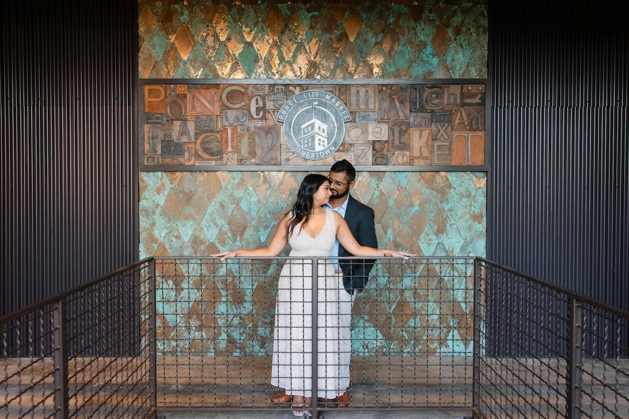 A couple stands behind a metal railing on an indoor balcony, with a decorative wall featuring a circular logo and colorful geometric patterns in the background.