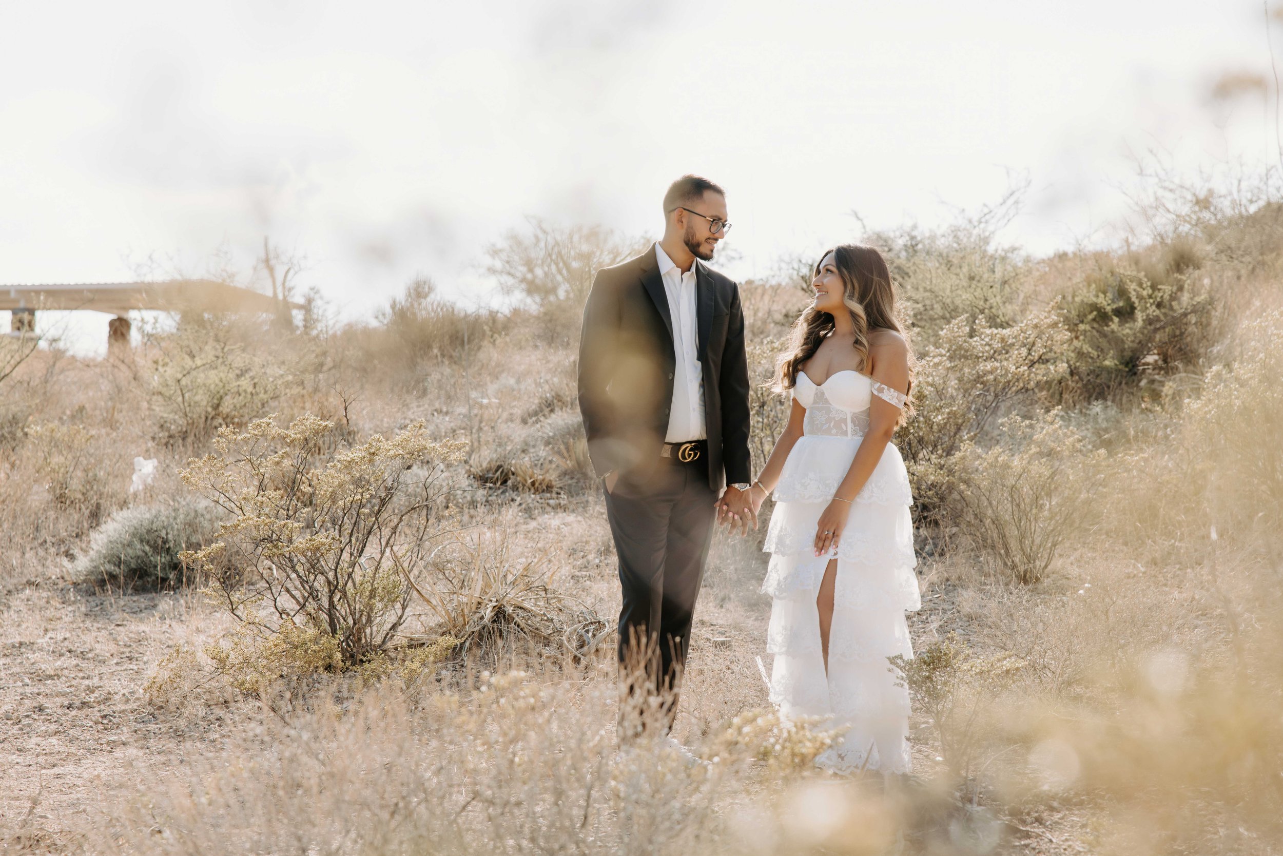 A bride and groom holding hands in a desert landscape, smiling at each other.